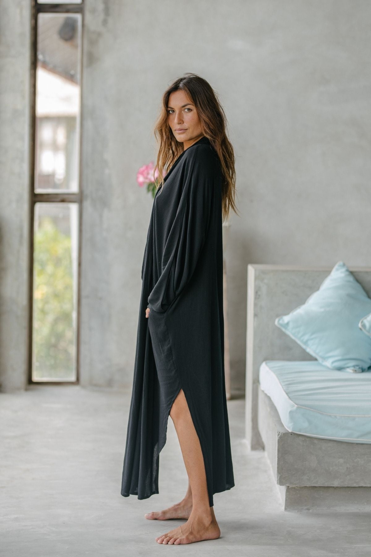 A woman stands barefoot indoors in an Amma Gown - Dark Moon Edition by Myrah Penaloza. She exudes intentional living in a minimalist room, featuring a concrete couch with blue cushions and a tall window, evoking the essence of an AMMA sanctuary.