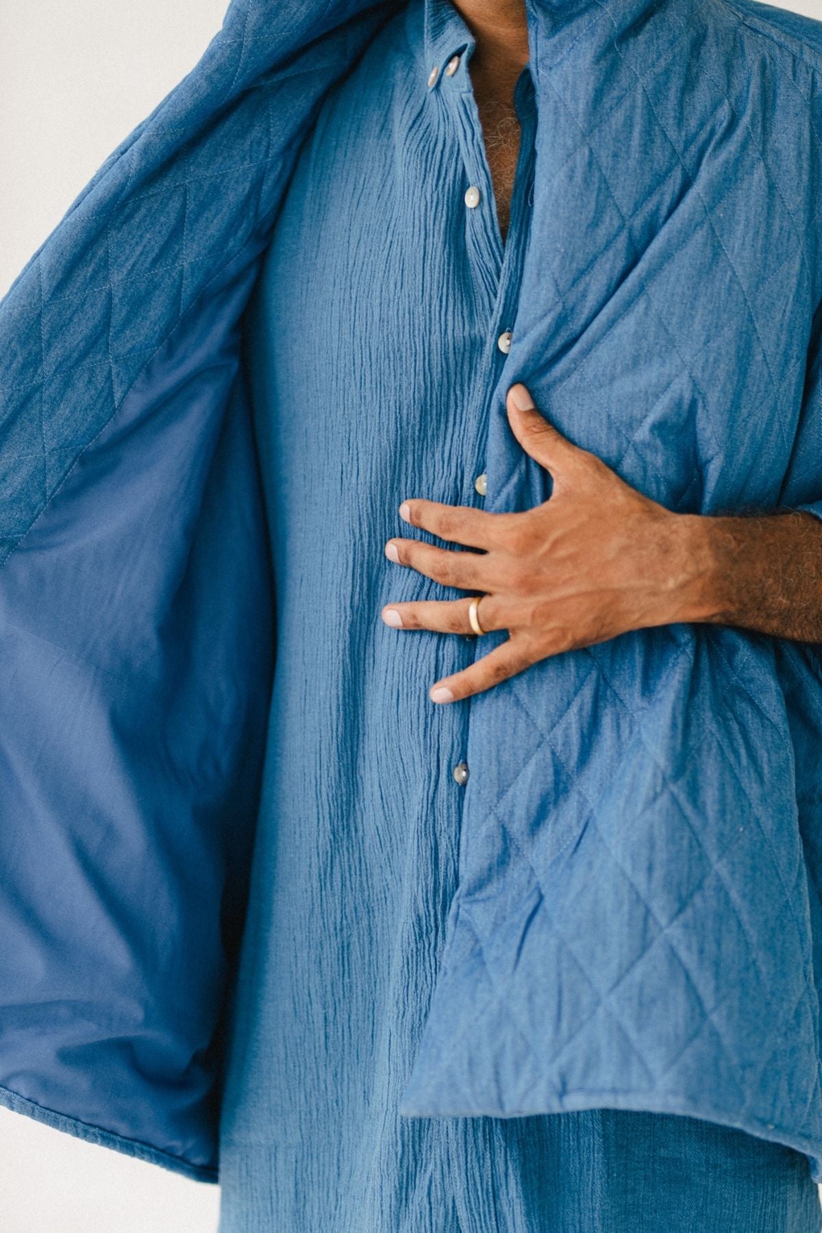 A person partly obscured by a Javi Quilted Jacket (Pre-Order) from Myrah Penaloza, draped over their shoulders, is wearing a textured blue shirt with buttons. They are holding the edge of the jacket with one hand, revealing the intricate inside lining.