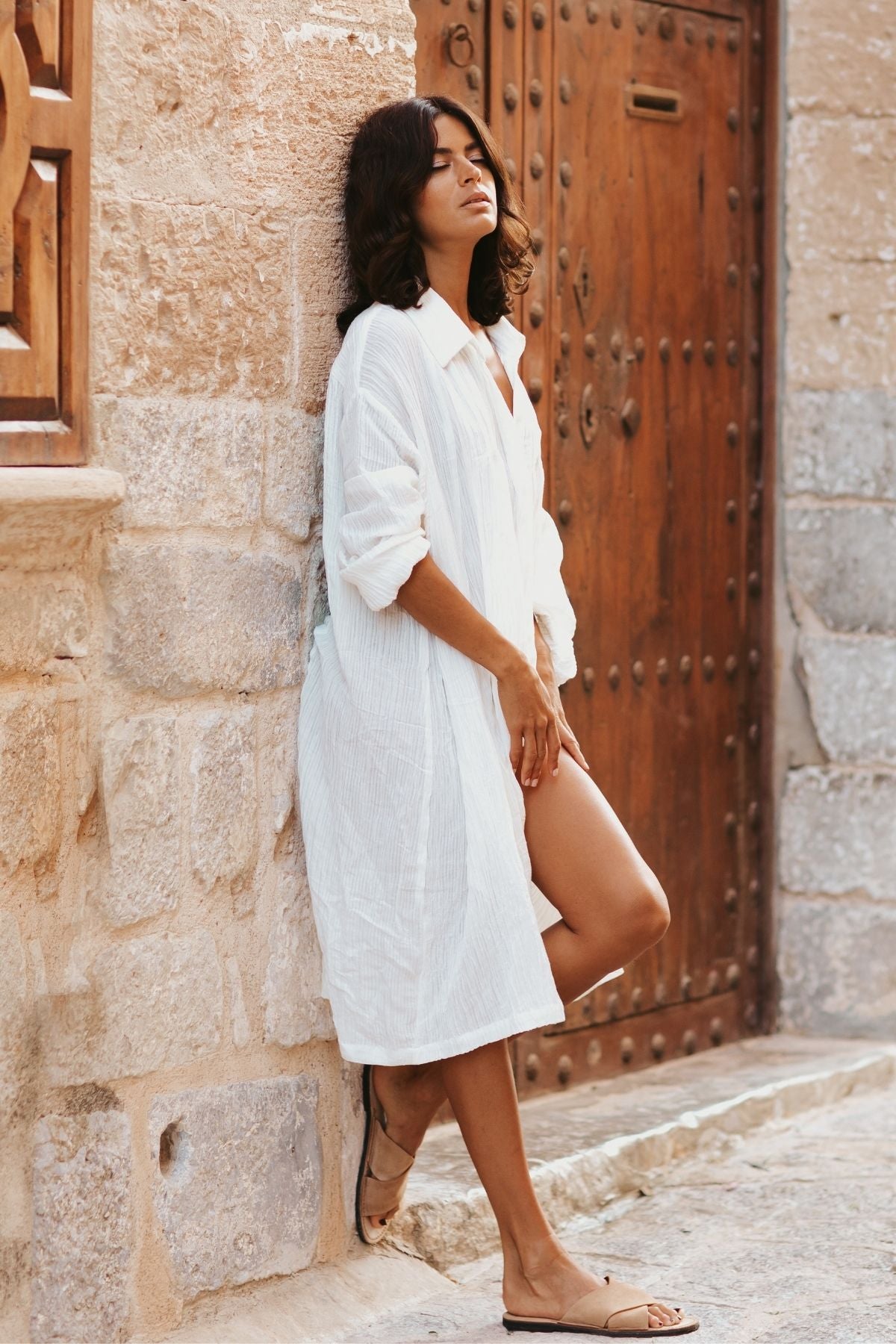 A woman in a Kundalini Gown - Short Edition from Myrah Penaloza, made of Linen/Cotton Luxe and white in color, leans against a stone wall with her eyes closed and head tilted back. She stands in front of a rustic wooden door, wearing beige sandals. The setting exudes old-world charm with its stone architecture.