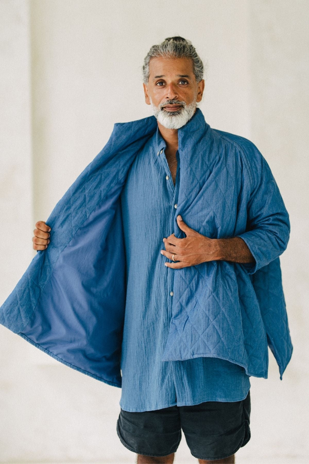 A middle-aged man with gray hair and a beard stands against a light background. He is wearing a Myrah Penaloza Javi Quilted Jacket (Pre-Order) in blue chambray denim over a blue shirt, holding the jacket open to reveal its hand-quilted inside. He also wears black shorts, and his expression is serious.