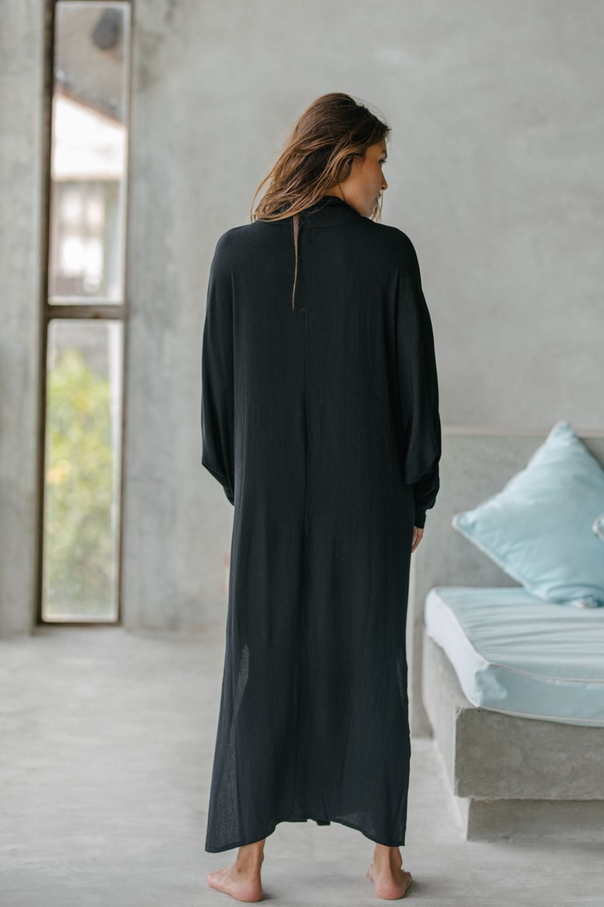 A woman with long hair stands barefoot indoors, wearing the Amma Gown - Dark Moon Edition by Myrah Penaloza, embracing intentional living. She faces away near a window and a gray bench with light blue pillows in an AMMA-inspired minimalist concrete interior.