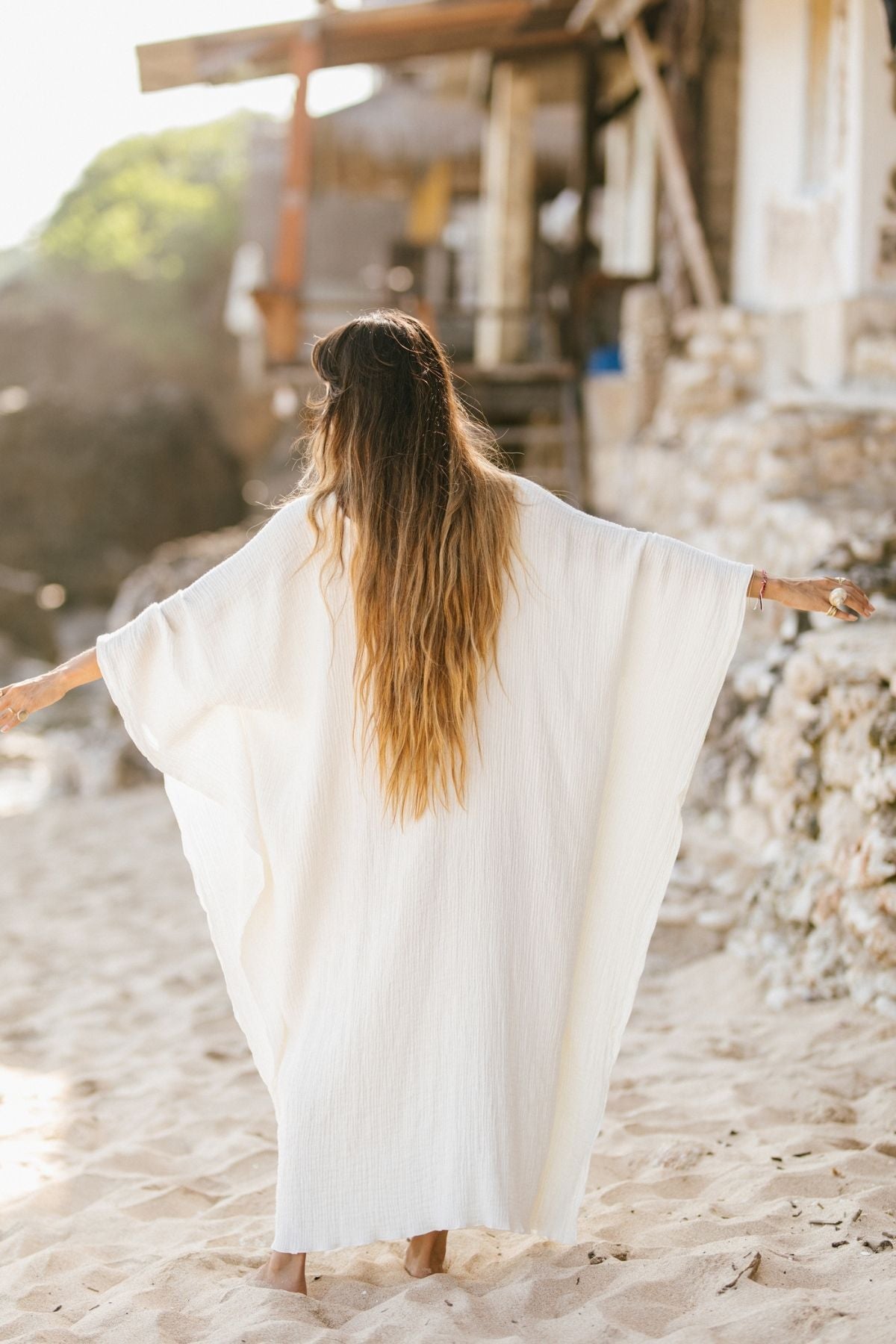 A person with long hair stands on a sandy beach, facing away from the camera while wearing the River Kaftan, a made-to-order piece crafted from 100% organic hemp and cotton by Myrah Penaloza. Their arms are outstretched near rustic buildings and rocks in the background.