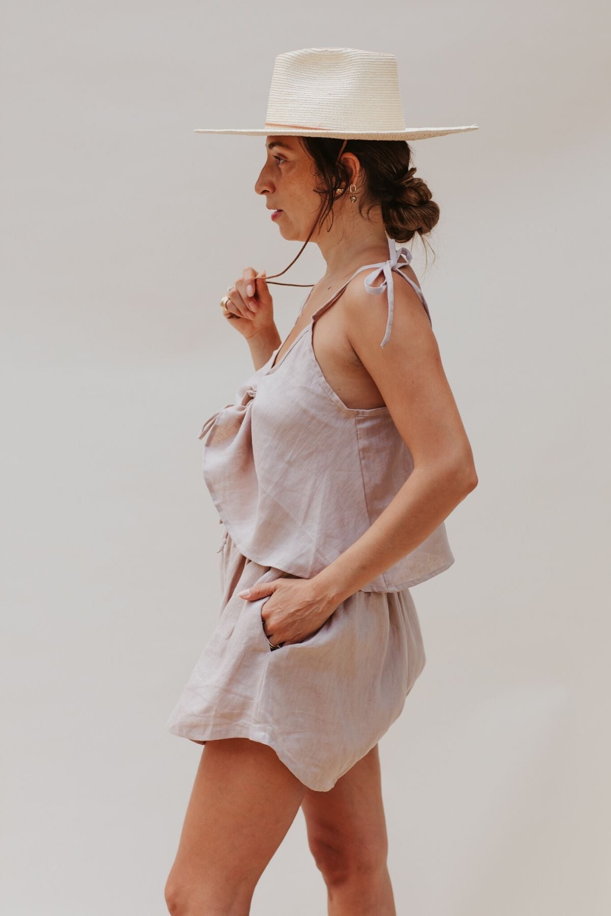 A woman in a Nidra Sleeper Top by Myrah Penaloza, made from 100% linen in clay, stands in profile against a plain background. She adjusts the strap of her sleeveless top while wearing a straw hat, with one hand resting in the pocket of her matching shorts. Her hair is styled in a low bun.