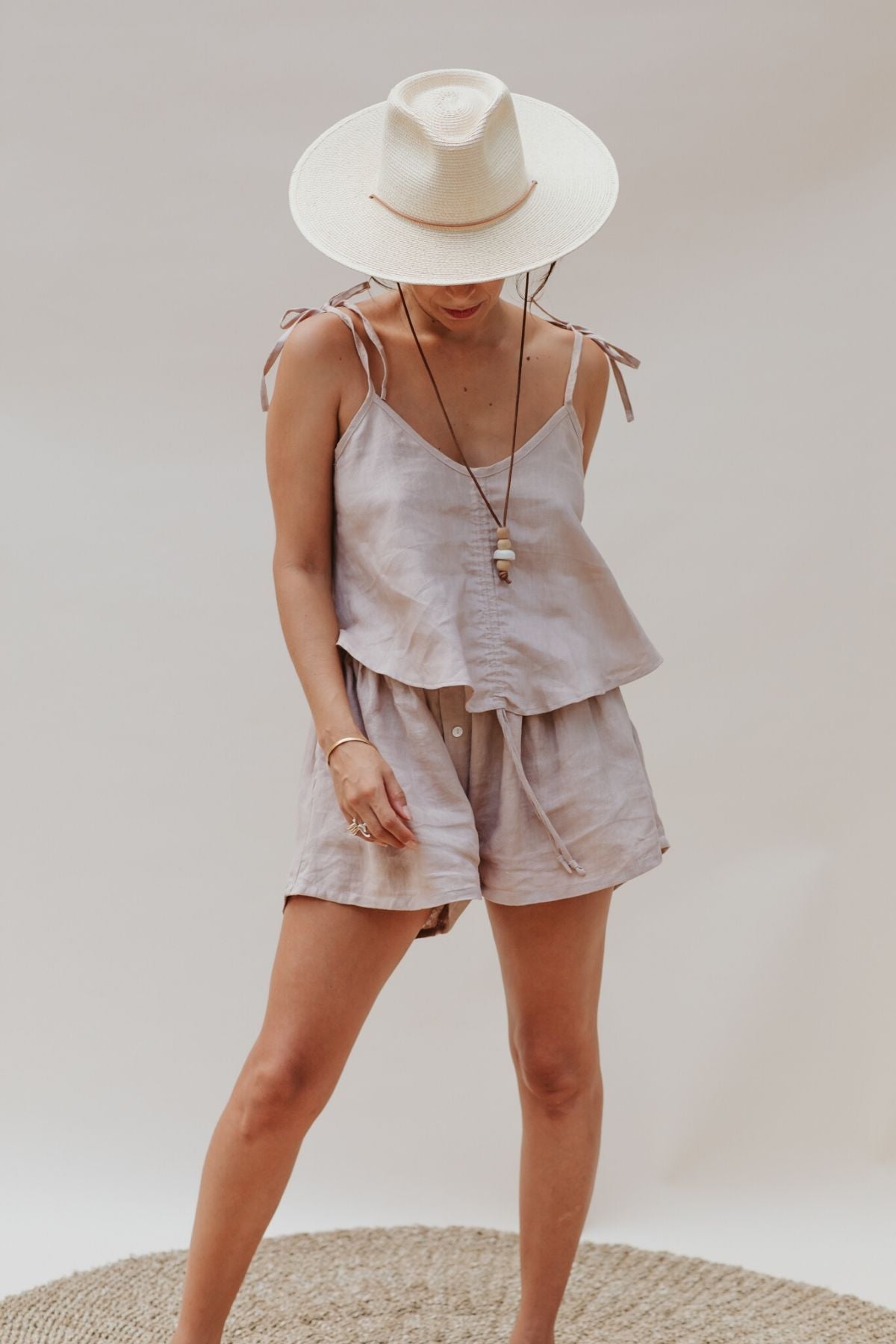 A person wearing a wide-brimmed hat and the Nidra Sleeper Top Sleeper in 100% linen, clay color by Myrah Penaloza, paired with matching shorts, stands on a woven rug. They are gazing down with their hands in their pockets against a simple, neutral backdrop.