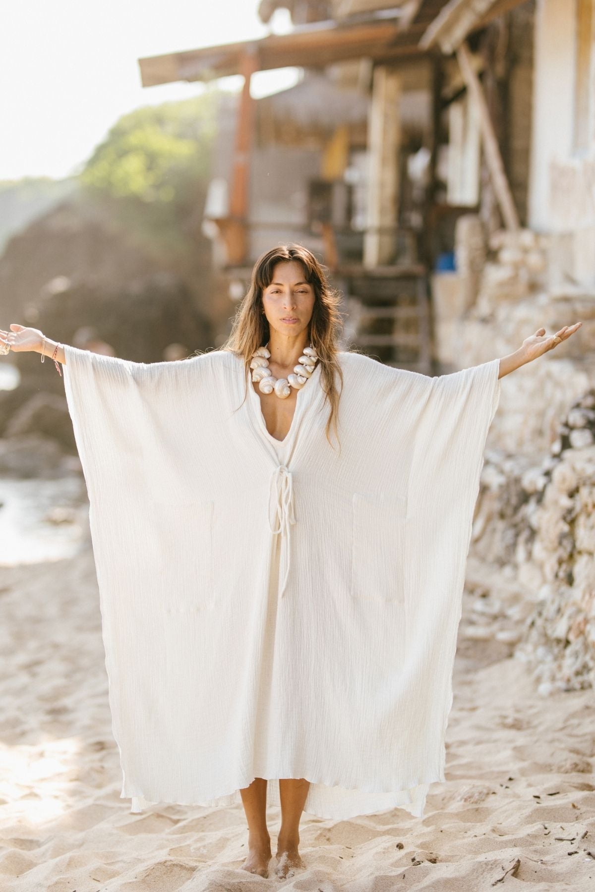 On a sandy beach, an individual raises their arms wide, adorned in a long, flowing River Kaftan crafted from 100% organic hemp and cotton by Myrah Penaloza, complemented by a statement necklace. A rustic wooden structure and lush greenery create a picturesque background.