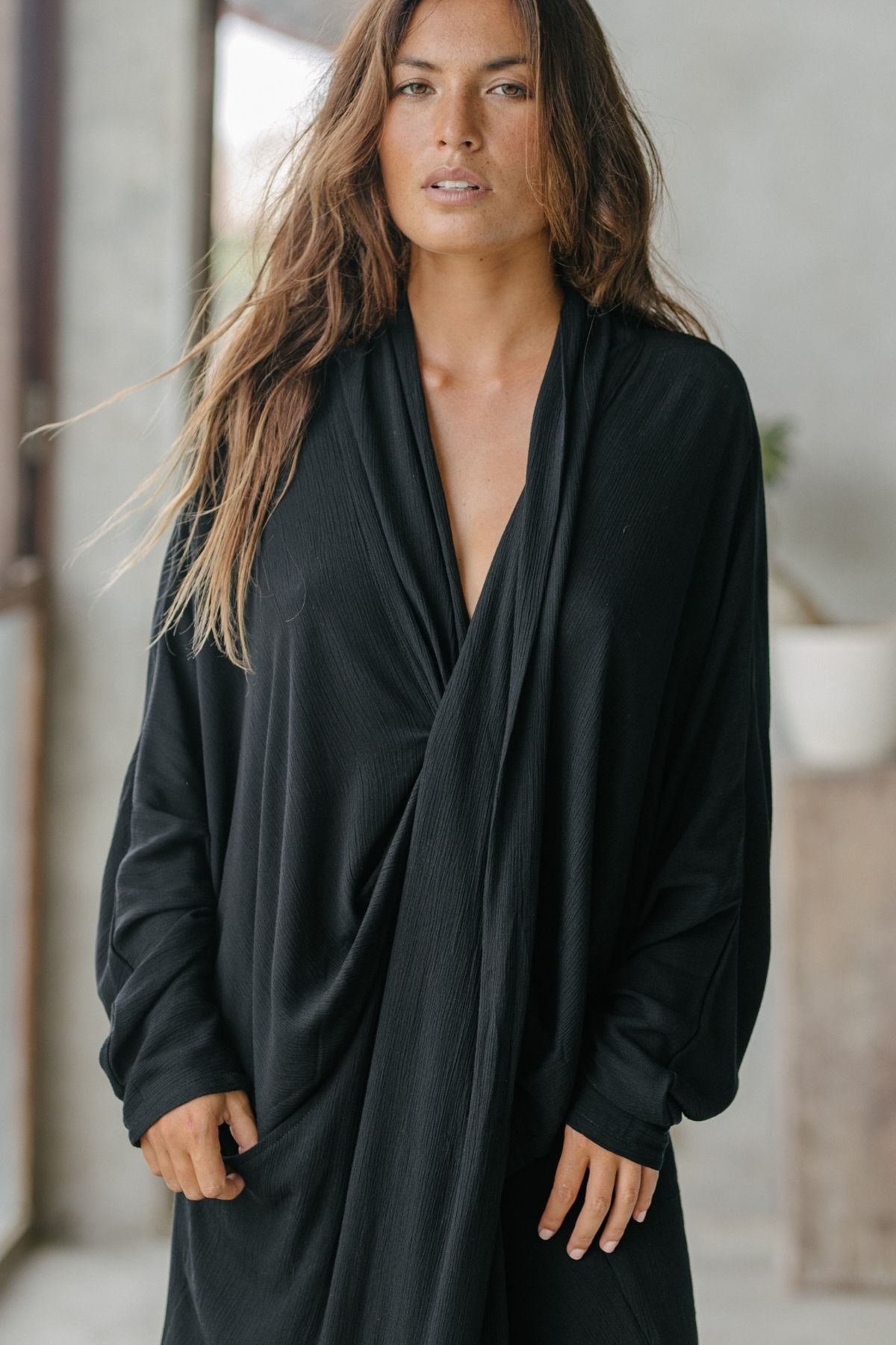 With long hair, a person embodying intentional living wears the Myrah Penaloza Amma Gown - Dark Moon Edition in bamboo rayon. They stand indoors, looking calmly at the camera against a softly blurred, minimalistic backdrop.