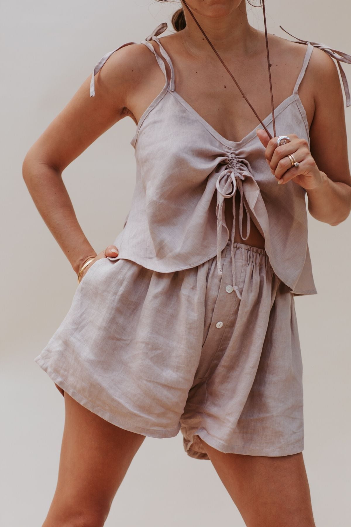 A person wearing a Nidra Sleeper Top by Myrah Penaloza, made from 100% linen in a clay hue, styled with complementary high-waisted shorts stands against a plain background. They hold sunglasses in one hand and rest the other on their hip, exuding casual sophistication.