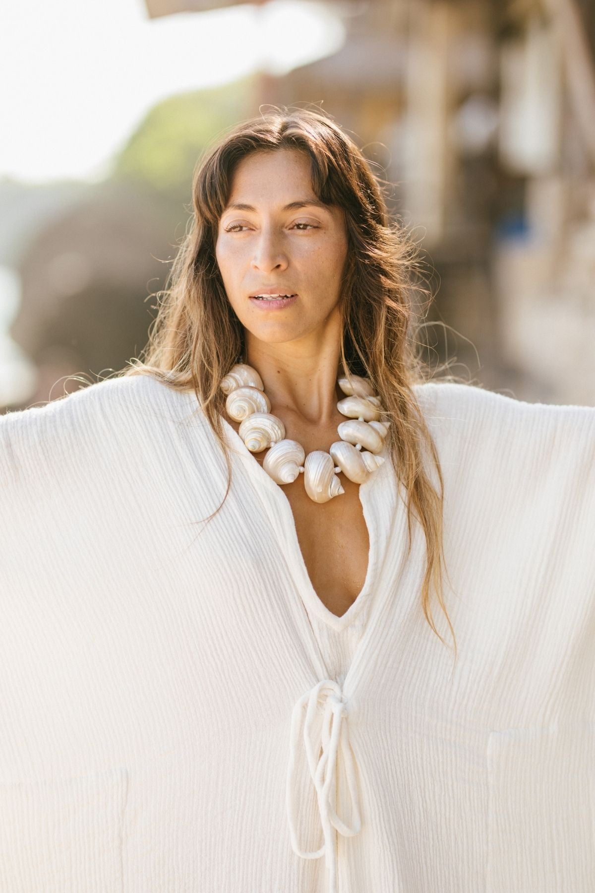 A woman is outdoors wearing the River Kaftan from Myrah Penaloza, crafted from 100% organic hemp and cotton, featuring a tie-front design and textured white fabric. She accessorizes with a substantial necklace made of white shells, while her long hair flows over her shoulders against a softly blurred natural backdrop.