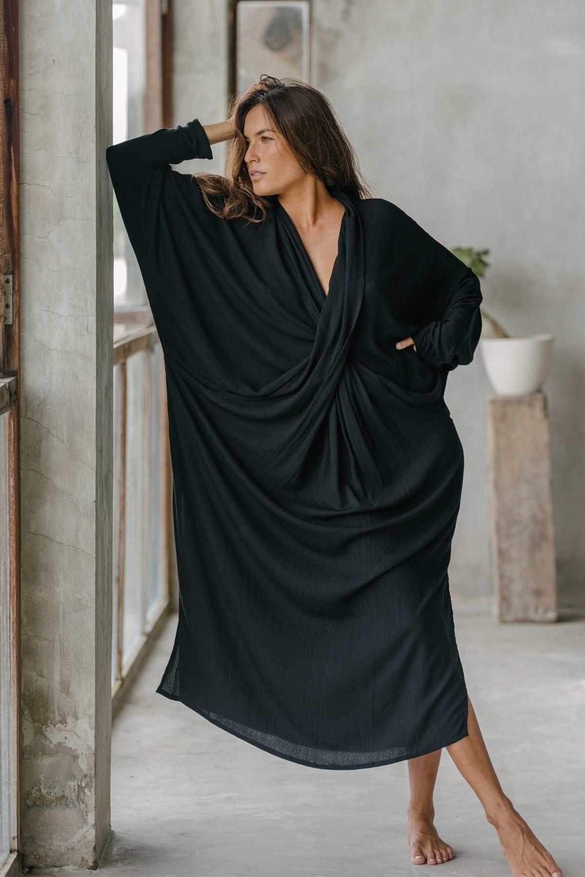 A woman in the Myrah Penaloza Amma Gown - Dark Moon Edition, crafted from bamboo rayon and evoking sacred elegance, stands by a large window. She leans gracefully against the wall, one arm raised, with her other hand in her pocket. A minimalist room accents intentional living with a potted plant.