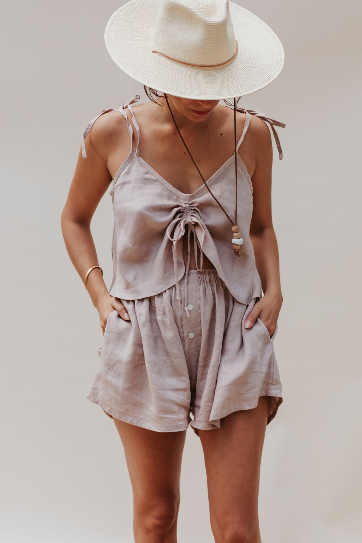 A person wearing a Nidra Sleeper Top by Myrah Penaloza, crafted from 100% linen in a clay hue with adjustable straps, paired with shorts and topped off with a wide-brimmed hat. The casual attire in soft neutral tones offers linen-like comfort, creating an effortlessly relaxed summer look.