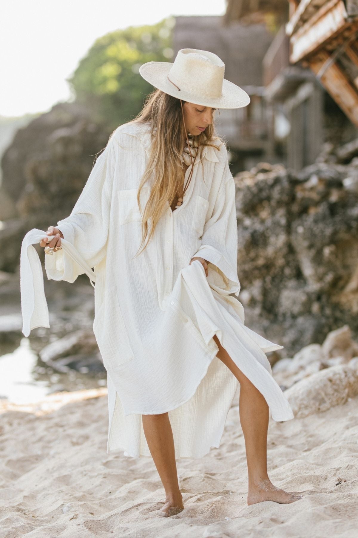 A woman stands barefoot on a sandy beach, wearing the Myrah Penaloza Nidra Duster, a flowy dress made of organic cotton and hemp. She holds the fabric and looks down, surrounded by rocky terrain and greenery, with a wide-brimmed hat complementing her look.