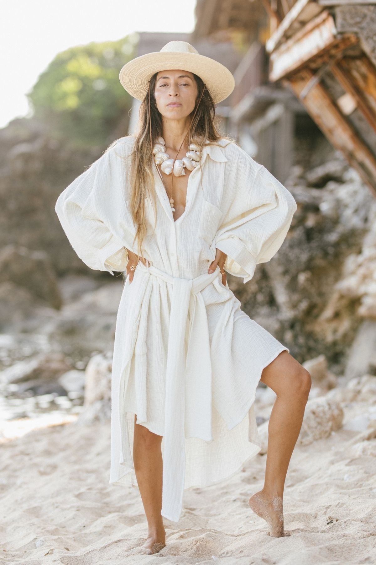 On a sandy beach, a person stands confidently wearing the Nidra Duster Organic Cotton/Hemp by Myrah Penaloza, styled in an oversized fit. The outfit is completed with a wide-brimmed hat and a large necklace. The backdrop of rocks, a wooden structure, and distant greenery beautifully captures the spirit of sustainable fashion.