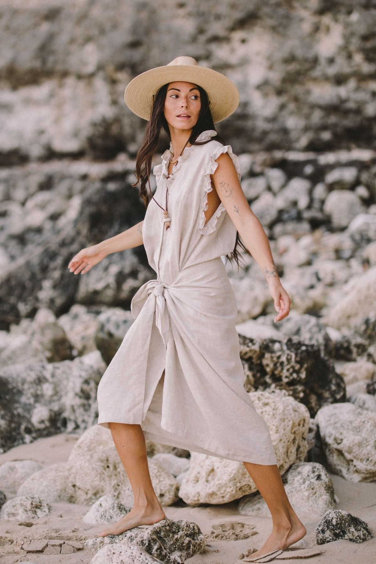 A woman strolls barefoot on a rocky beach, wearing The Madu Linen Wrap Dress from Myrah Penaloza and a wide-brimmed straw hat. Her long dark hair is adorned with ribbons, showcasing visible tattoos on her arms. The scene is set against large rocks and sand.