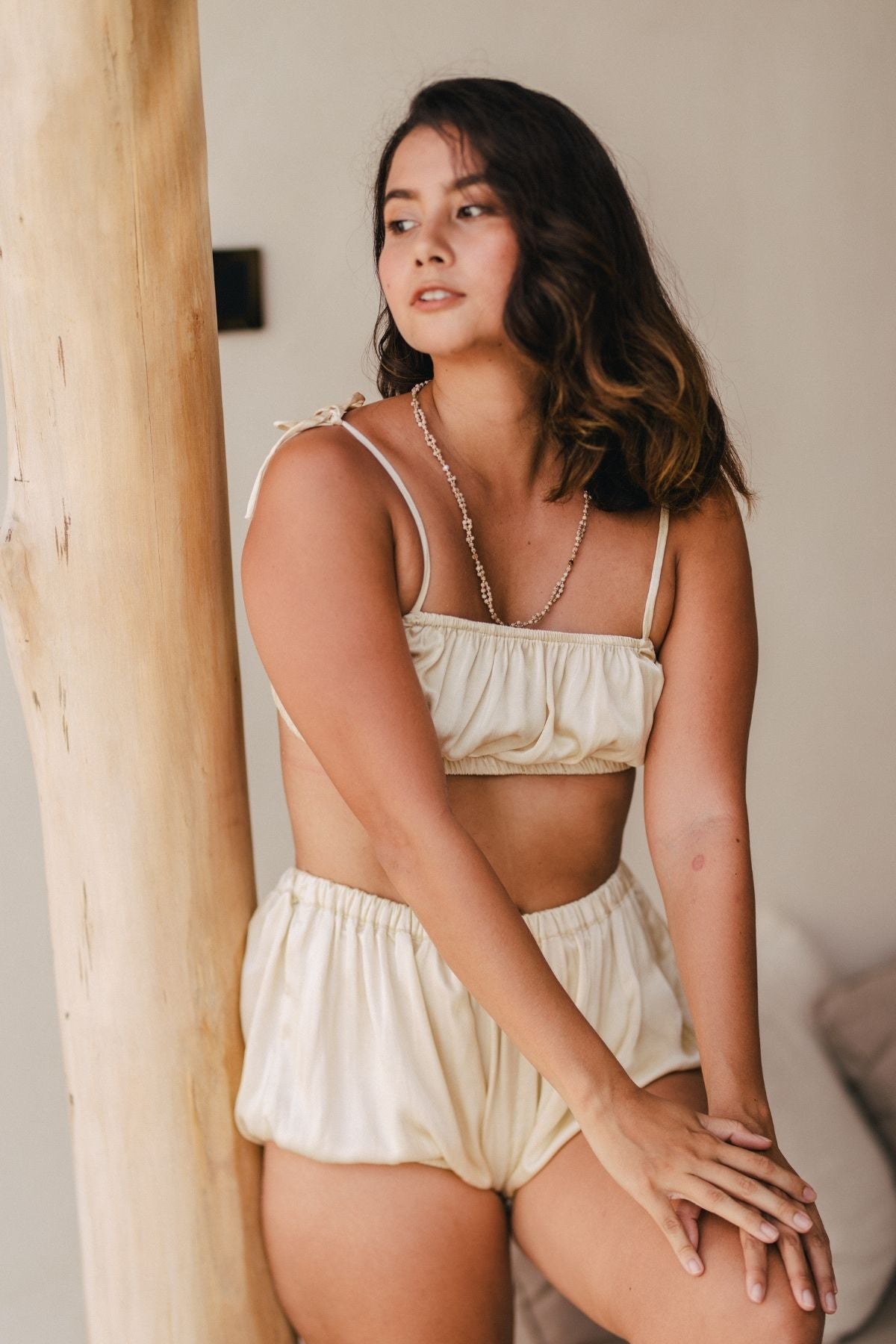 A woman with shoulder-length dark hair is leaning against a wooden beam, wearing a creamy-colored Lucid Dreamer Underwear Set in silk and satin by Myrah Penaloza. Bathed in moonlight, she looks off to the side with her hands resting on the beam and a relaxed, contemplative expression.