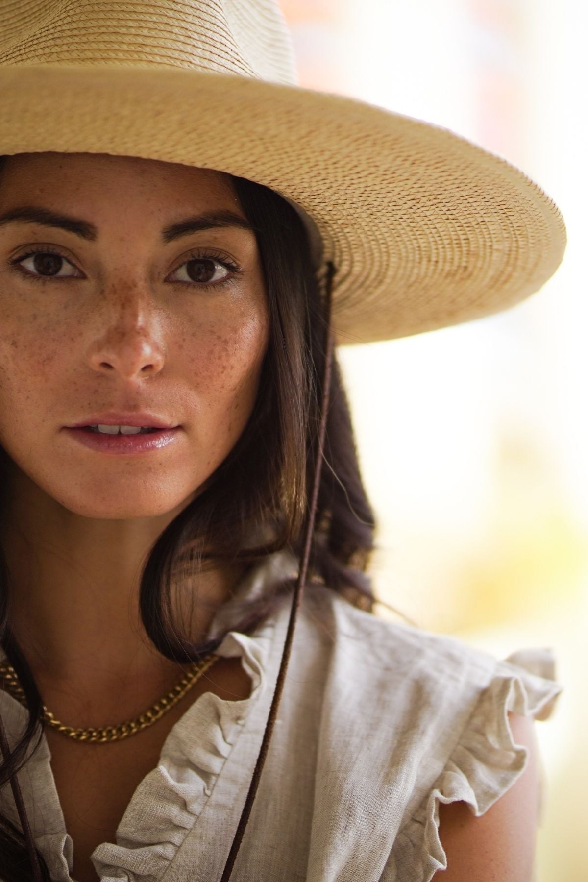 A woman with long dark hair and freckles is wearing The Madu Linen Wrap Dress by Myrah Penaloza, which features a wide-brimmed straw hat and a cream-colored design with ruffled sleeves. She gazes directly at the camera against a softly blurred background, creating a warm, serene atmosphere.