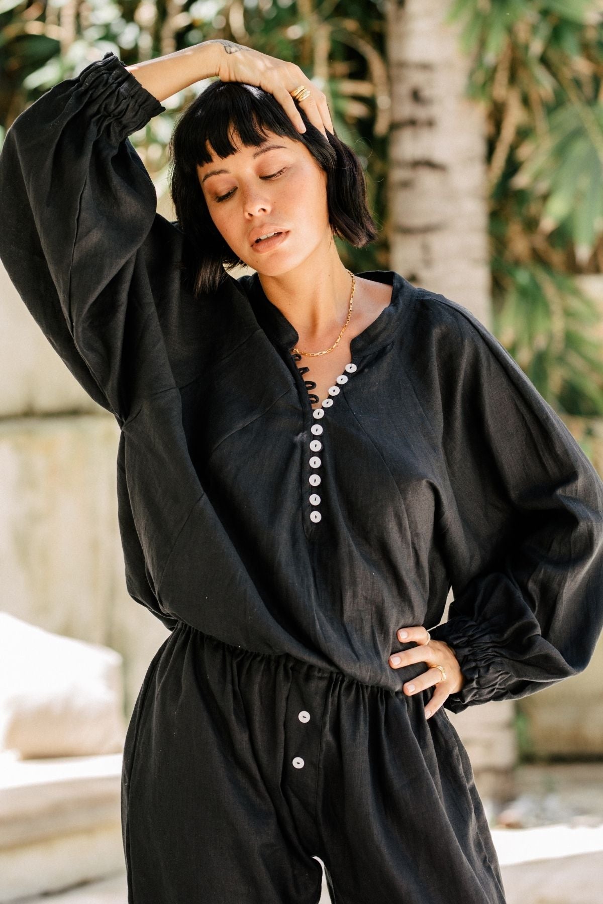 A person with short dark hair is posing outdoors, wearing the black, long-sleeve Frida Top from Myrah Penaloza, made from 100% linen and featuring white buttons on the top and bottom. The individual has one hand resting on their head and the other on their hip, with green leaves and a tree in the blurred background.