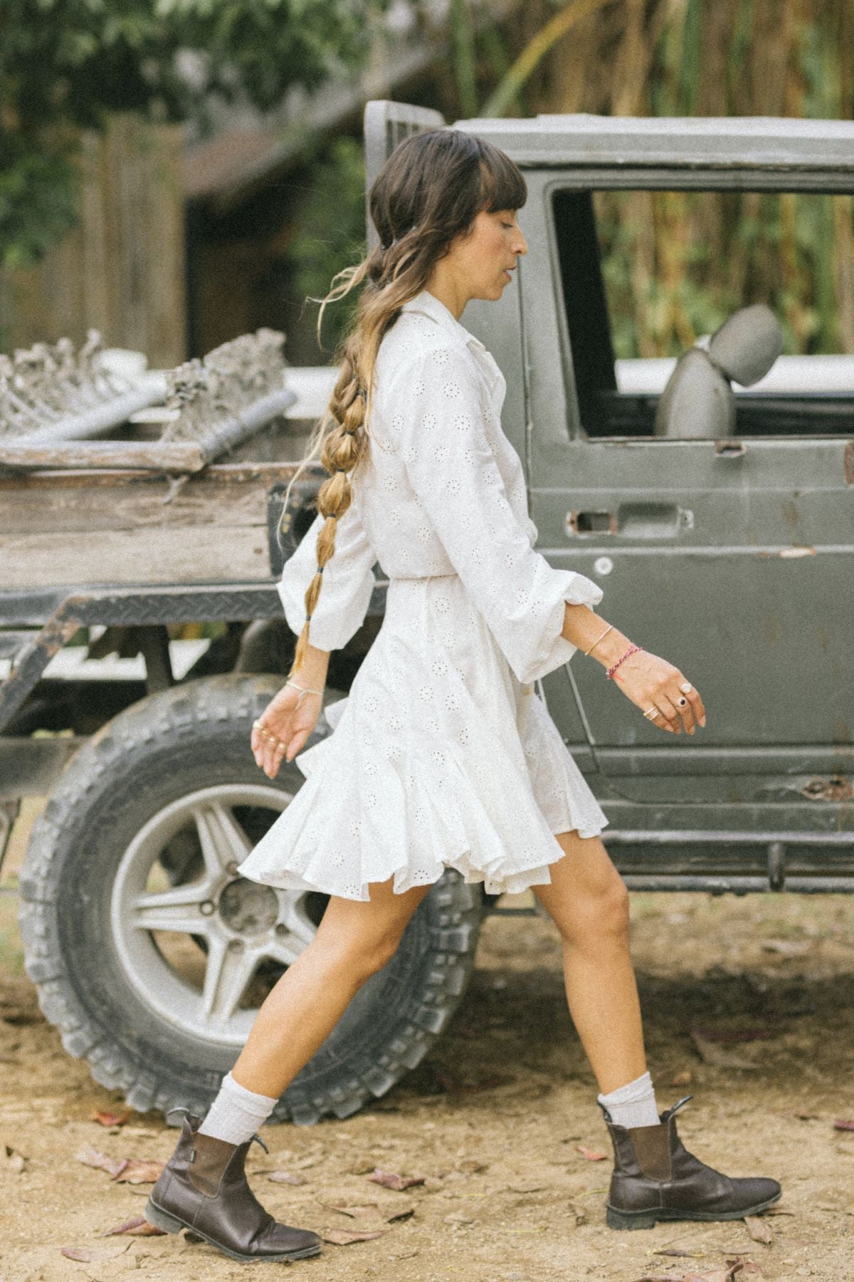 A woman in a La Rosa Dress by Myrah Penaloza, featuring vintage embroidered cotton, with a long braid strolls next to a rugged vehicle on a dirt path. She wears brown ankle boots and is encircled by lush greenery. The flatbed holds various items, suggesting an adventurous journey with carbon neutral shipping in consideration.