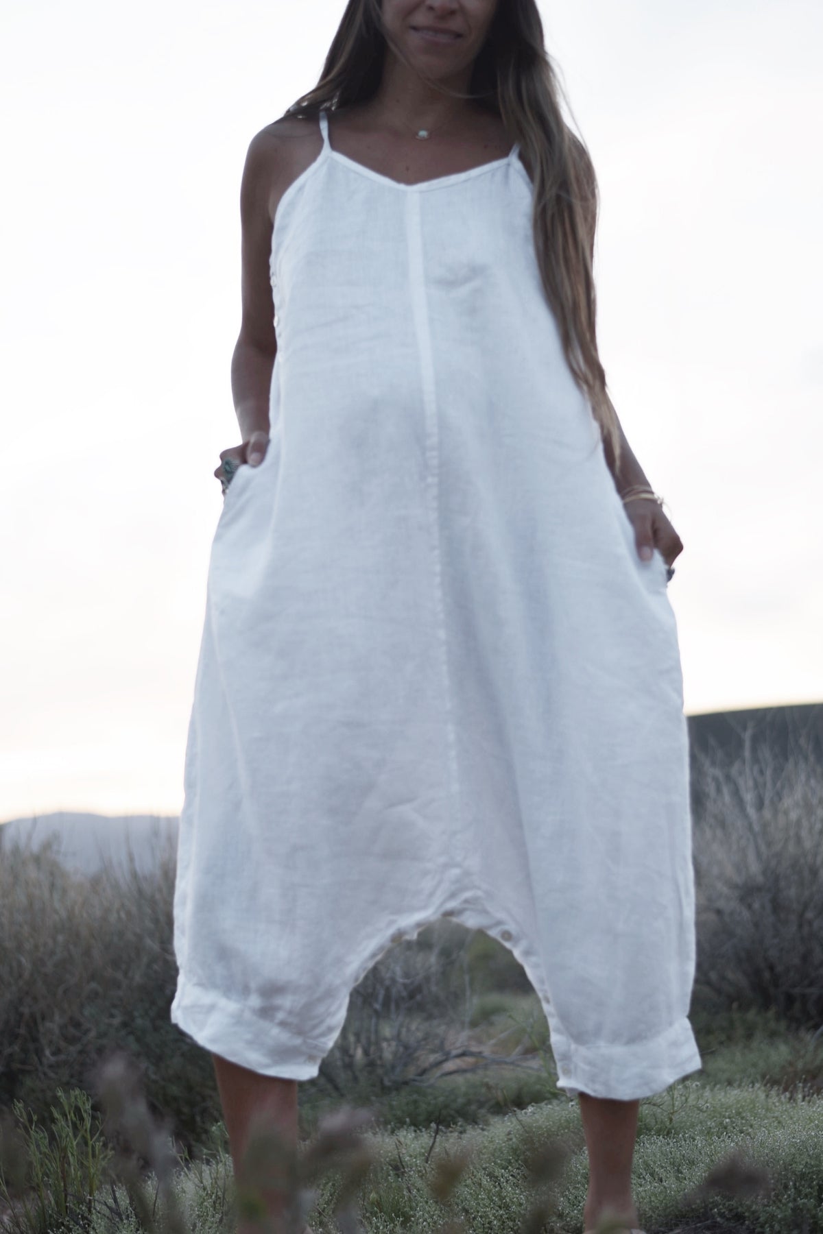 A woman in a white, loose-fitting Japa Playsuit from Myrah Penaloza stands outdoors. The Japa Playsuit, made from 100% breathable linen fabric, is sleeveless with thin straps and features a harem-style silhouette that ends at her mid-calf. She has long hair and is surrounded by grass and bushes in the natural setting.