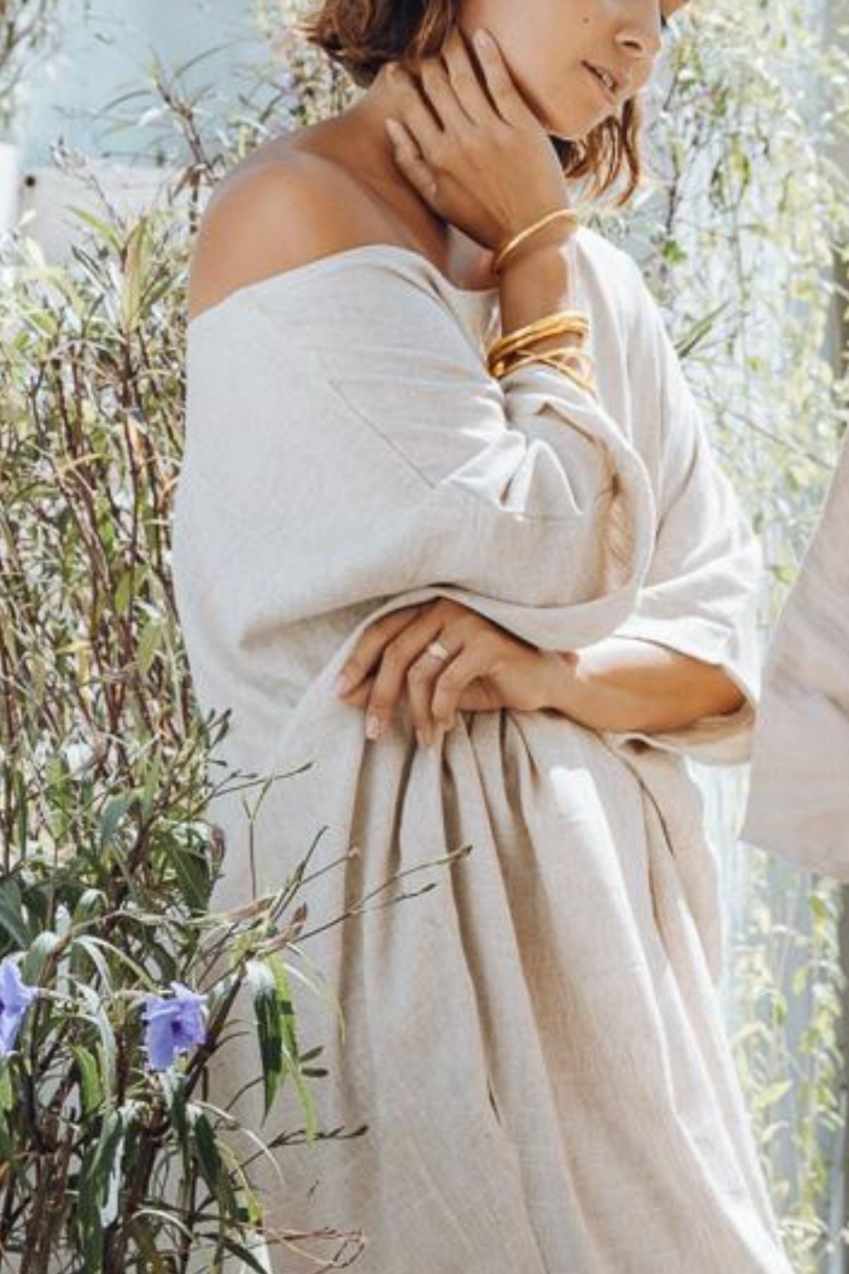 A woman wearing the Butterfly Tunic by Myrah Penaloza stands amidst greenery, the loose linen falling elegantly off her shoulders. She accessorizes with gold bangles, gently placing her hands near her face and waist. Purple flowers bloom in the foreground, adding charm to this everyday piece.