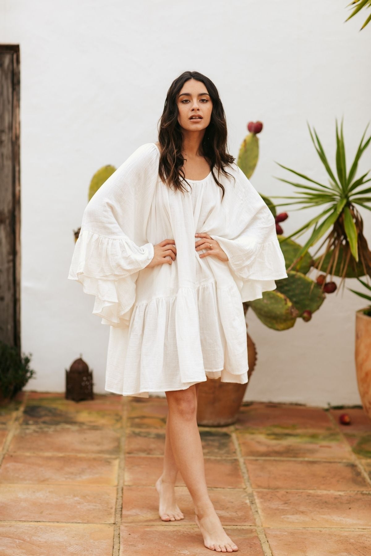 A barefoot woman in a flowing, white Brida Gown Short (WI) by Myrah Penaloza stands on a terracotta-tiled patio. Behind her are various plants, including a spiky green succulent and a cactus with red blooms. She gazes directly at the camera with a serene expression as if inspired by Paulo Coelho's prose.