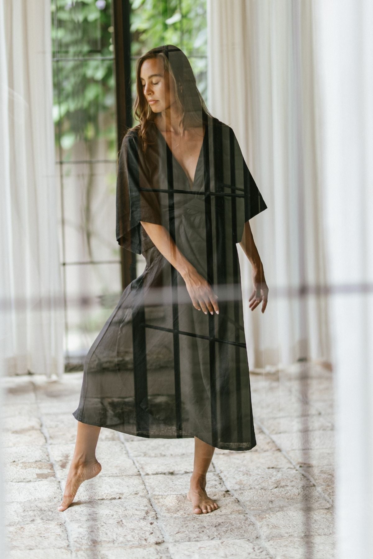 A woman wearing the Magdalena Gown by Myrah Penaloza, crafted from 100% linen in a moonlight hue, stands gracefully on tiptoe, posed in a dance-like movement. She is near a large window that lets in soft, natural light. The setting is airy and serene, with sheer curtains and a textured floor.