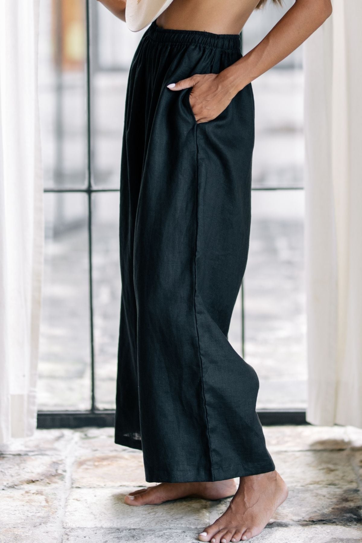 A person stands indoors near a window, dressed in elegant office wear featuring the stylish dark moon black Grace Pant made from 100% linen by Myrah Penaloza, paired with a light top. They are barefoot, with one hand tucked in a pocket, surrounded by sheer curtains and standing on a stone floor.