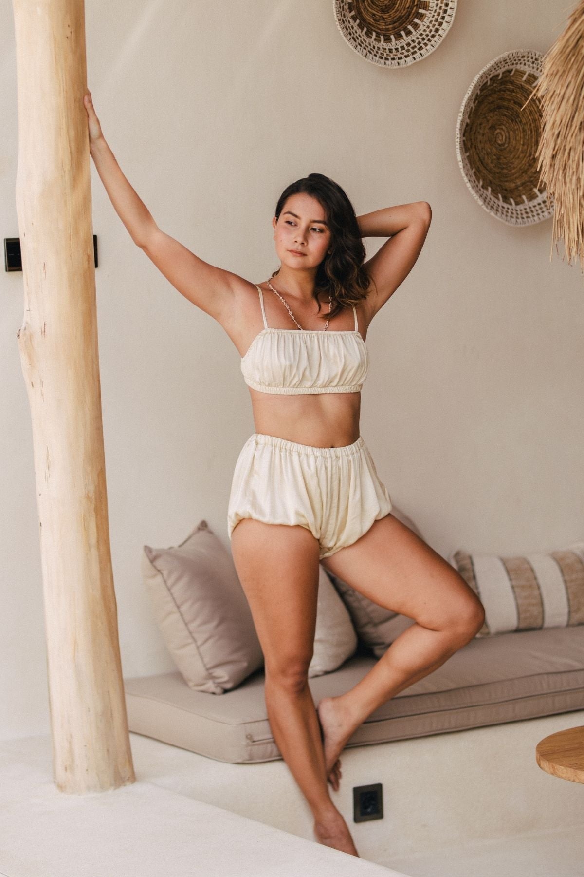 A woman stands against a wooden pole in a relaxed pose, wearing the Lucid Dreamer Underwear Set (Silk/Satin) - BGN by Myrah Penaloza. She has one arm resting on the pole and the other bent behind her head. The background consists of a cozy seating area with cushions and decorative woven wall hangings illuminated by moonlight.