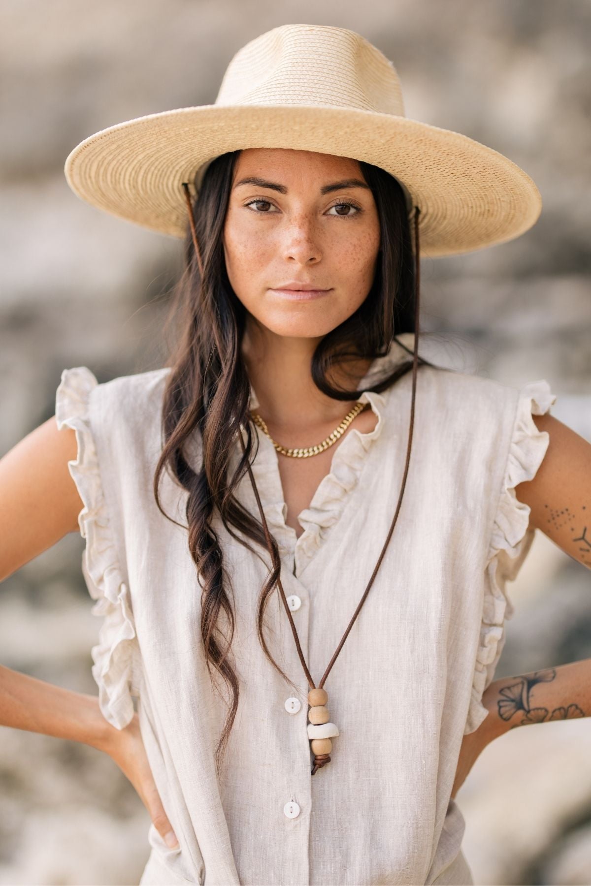 A person wearing The Madu Linen Wrap Dress by Myrah Penaloza, accessorized with a wide-brimmed straw hat and a necklace. Wrapped in chic style, they have long, dark hair and are standing outdoors with a serious expression. The background is blurred, emphasizing the individual.