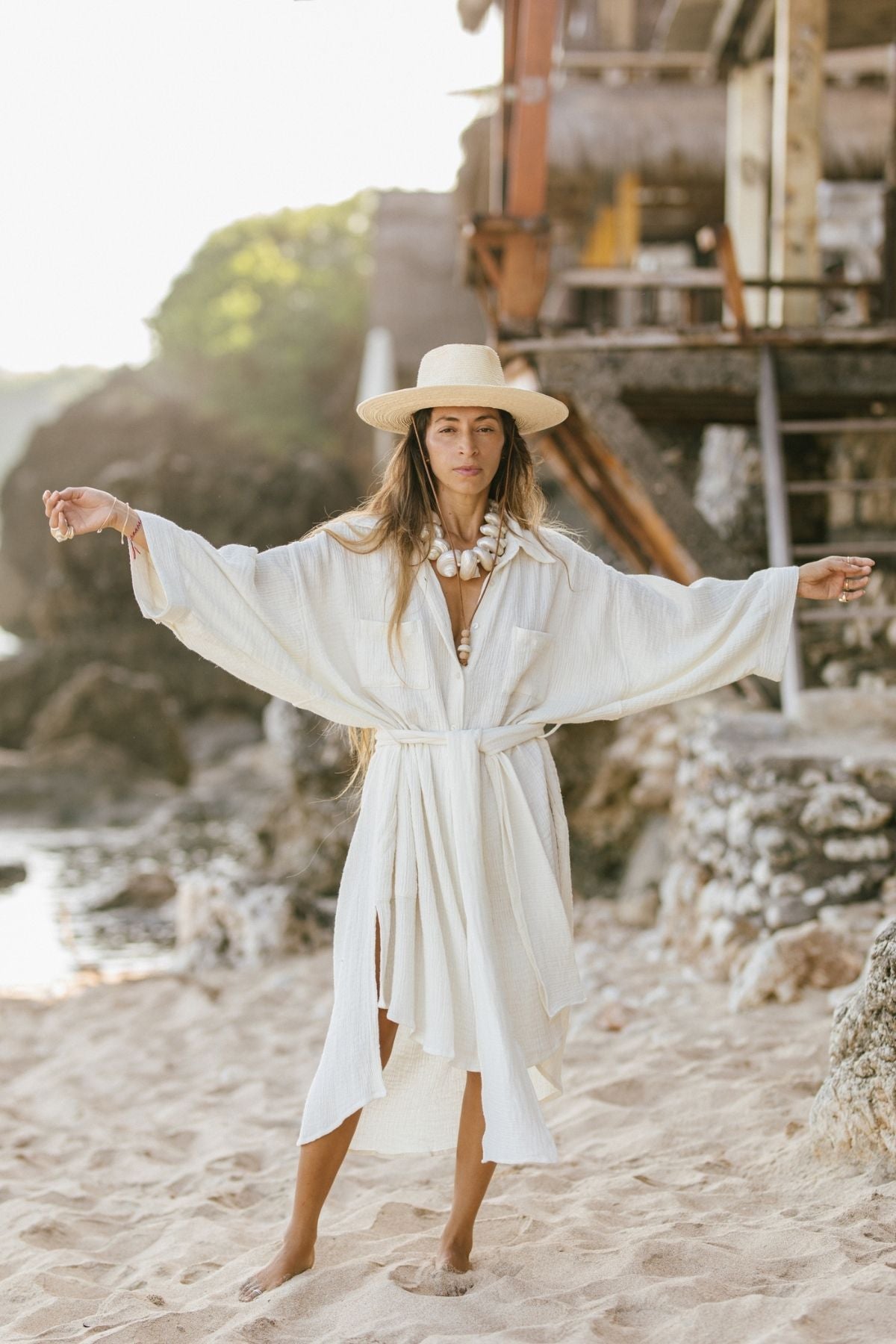 A person wearing the Nidra Duster from Myrah Penaloza, an oversized fit crafted from organic cotton and hemp, stands with outstretched arms on a sandy beach. They don a wide-brimmed hat and flowing white robe near rocks and ocean water, with a rustic wooden structure in the background.