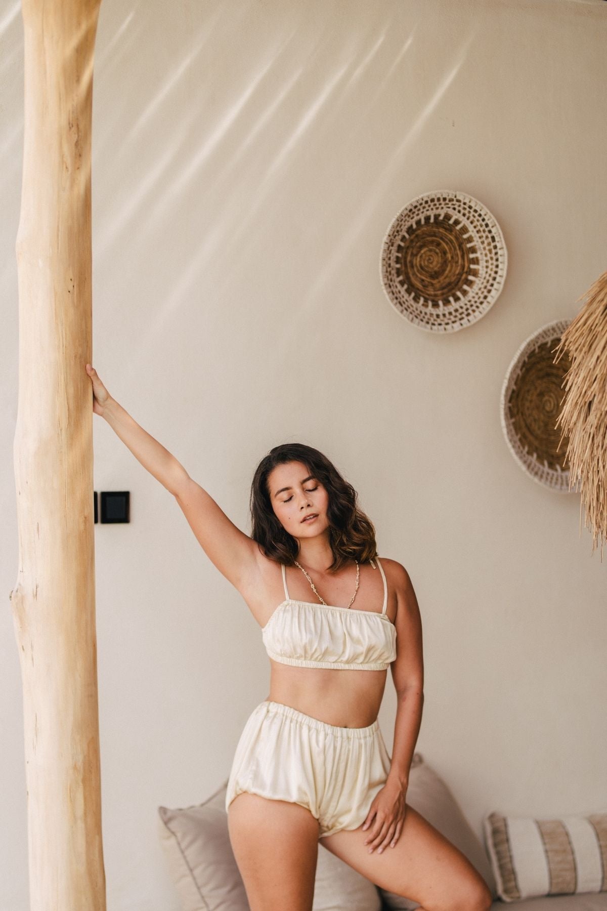 A woman stands indoors next to a wooden pillar, wearing the Lucid Dreamer Underwear Set (Silk/Satin) - BGN by Myrah Penaloza. The background features neutral-toned decor, including woven wall hangings and a thatched element on the right. She leans casually against the pillar with one arm raised, bathed in soft moonlight.