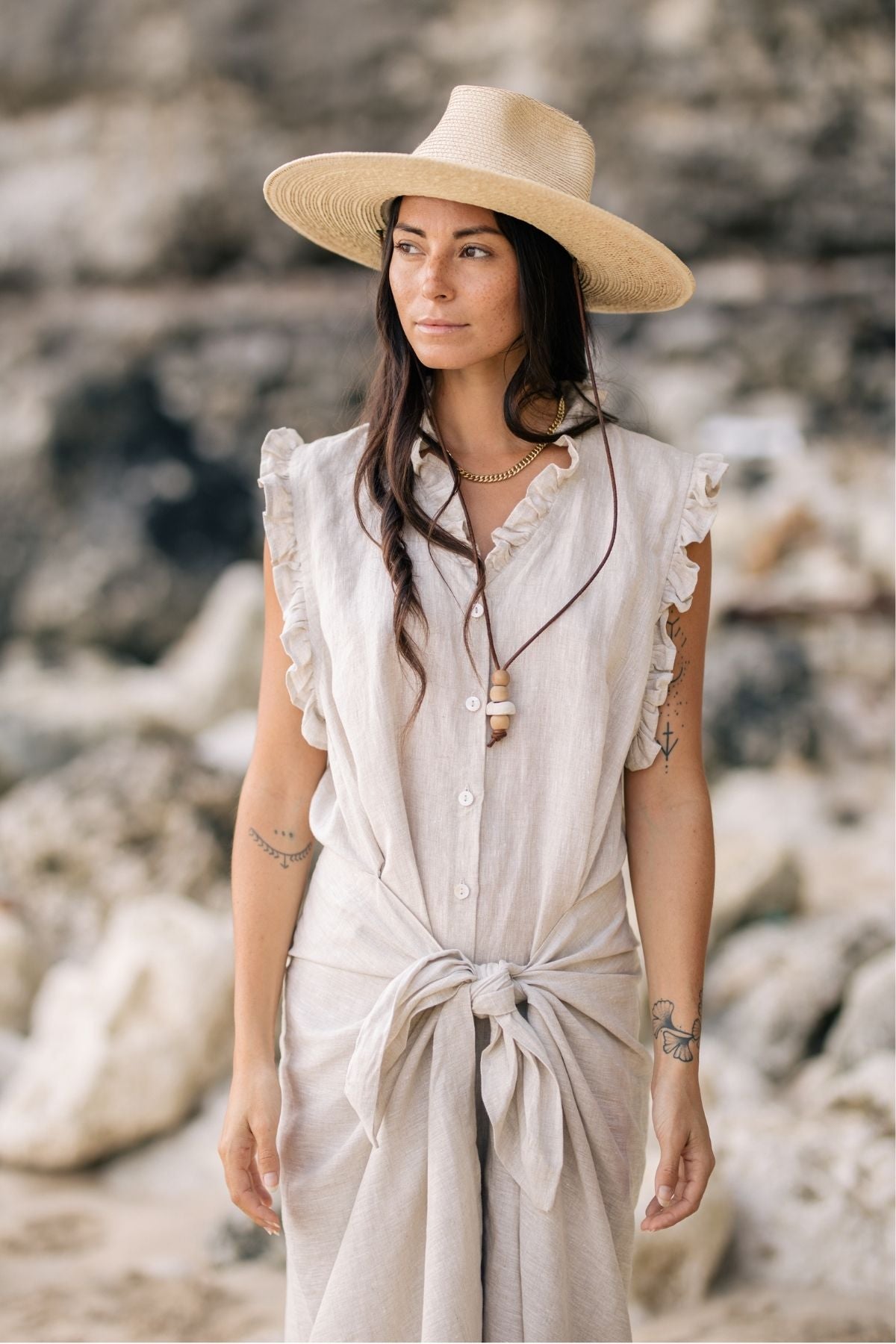 A woman wearing The Madu Linen Wrap Dress by Myrah Penaloza and a wide-brimmed straw hat stands on a rocky beach. She has long hair, tattoos on her arms, and wears a necklace. The wrap dress features ruffle details and a tie at the waist as she gazes into the distance.