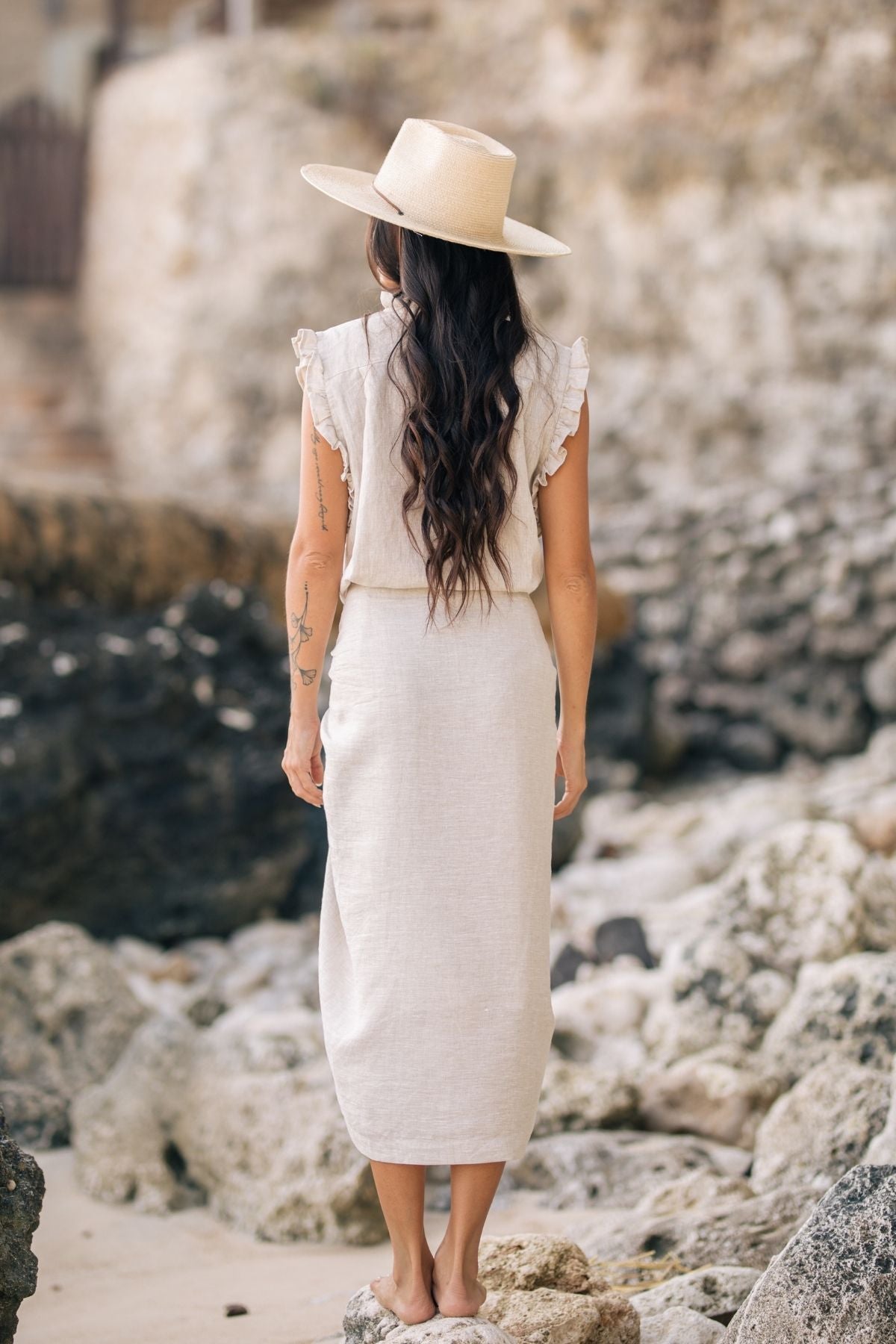 A woman with long hair stands on rocky terrain, facing away. She wears The Madu Linen Wrap Dress from Myrah Penaloza and a wide-brimmed hat. The background features stone and rough textures, creating a natural, rustic atmosphere.