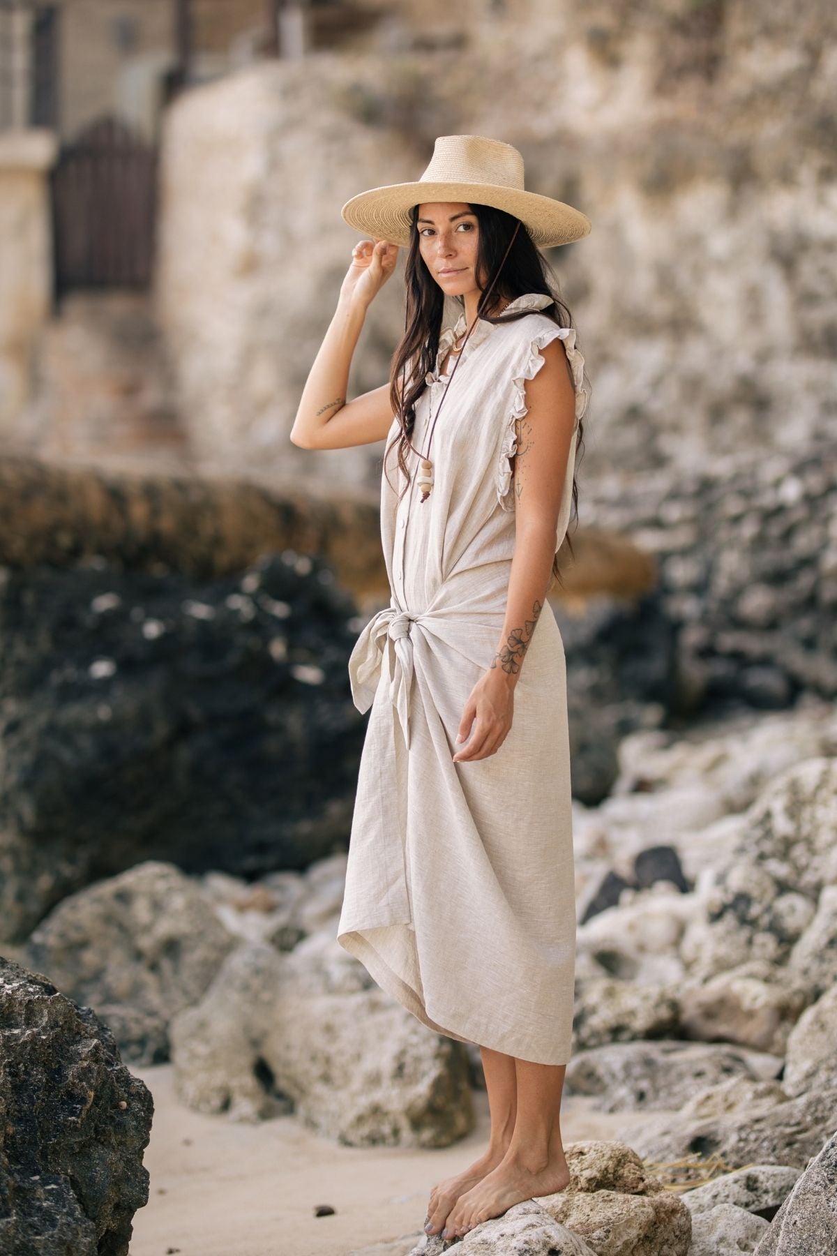 A woman stands barefoot on a rocky beach, wrapped in The Madu Linen Wrap Dress by Myrah Penaloza and a wide-brimmed hat. She has long dark hair and looks towards the camera, gently holding her hat. The background features rocks and a blurred structure.