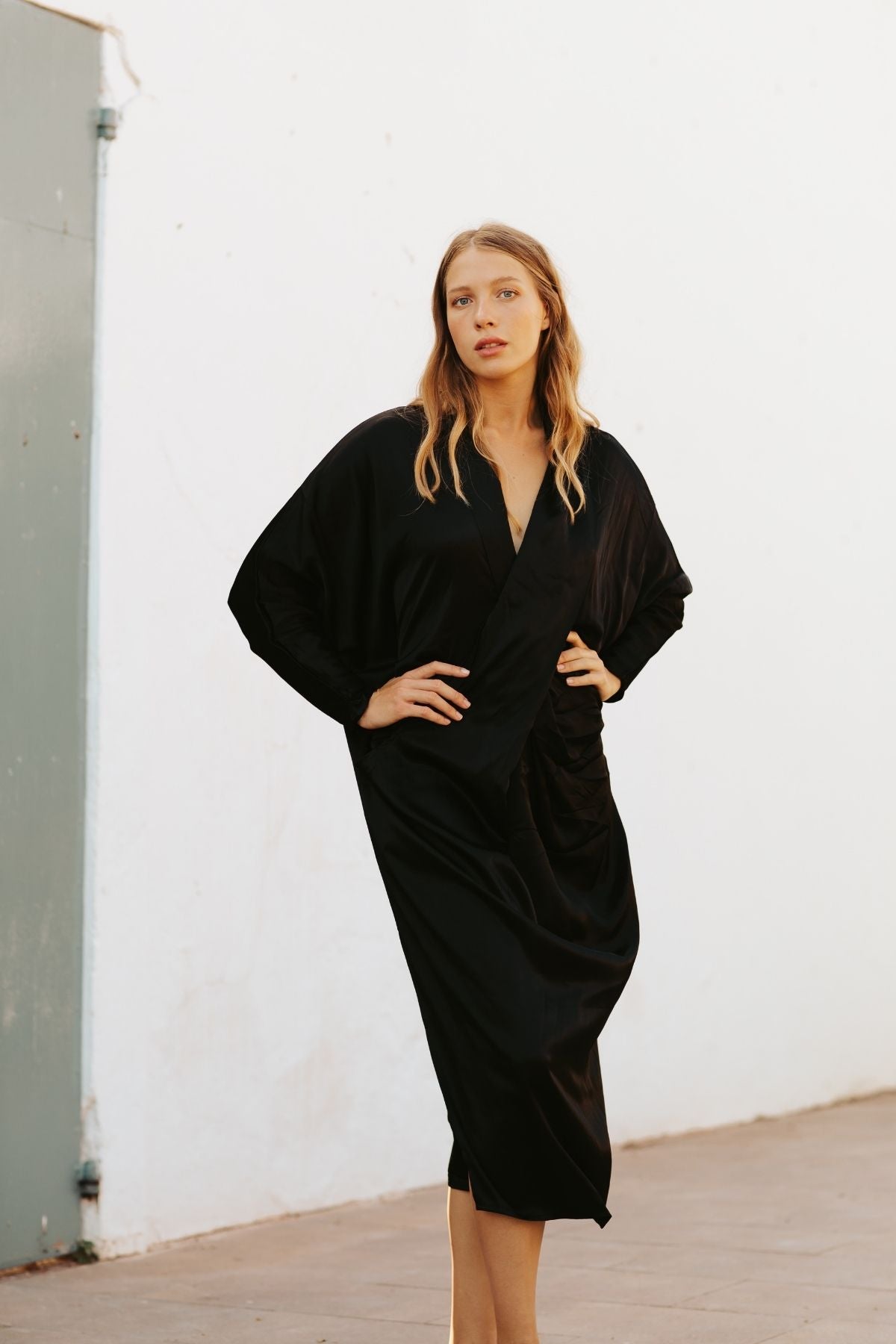 A woman stands against a white wall in the Amma Gown - Dark Moon Edition by Myrah Penaloza, exuding confidence and embracing intentional living. Her long blond hair flows as she poses with one hand on her hip outdoors, emphasizing the minimalist essence of this sacred bamboo rayon garment.