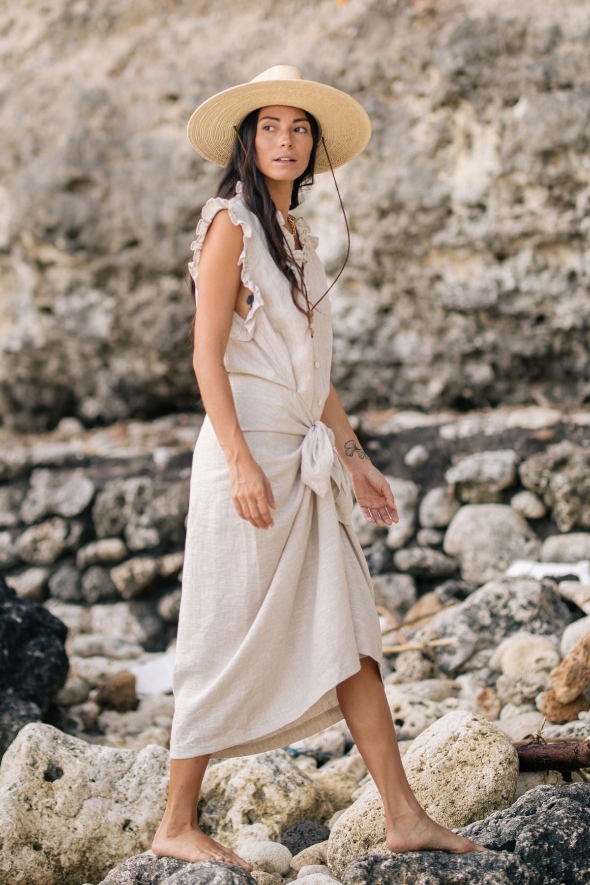 A woman is seen wearing The Madu Linen Wrap Dress by Myrah Penaloza paired with a wide-brimmed hat as she strolls barefoot across a rocky beach, with large textured stones and a natural, rugged landscape in the background.