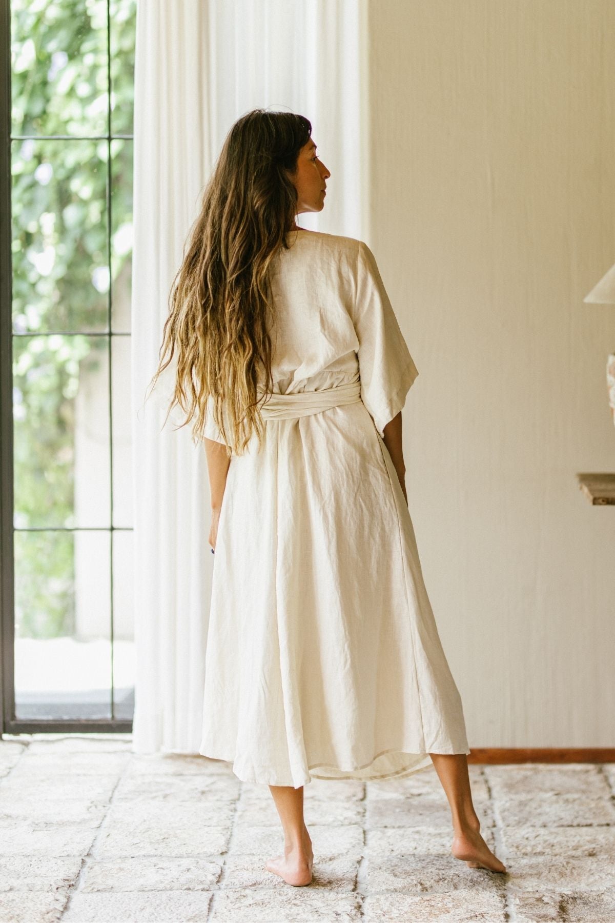 A woman with long hair stands barefoot in a bright, naturally lit room with a large window. She is wearing the Magdalena Gown by Myrah Penaloza, an adjustable wrap dress tied at the waist. The minimalist decor of the space complements her Moonlight-colored flax linen attire perfectly.