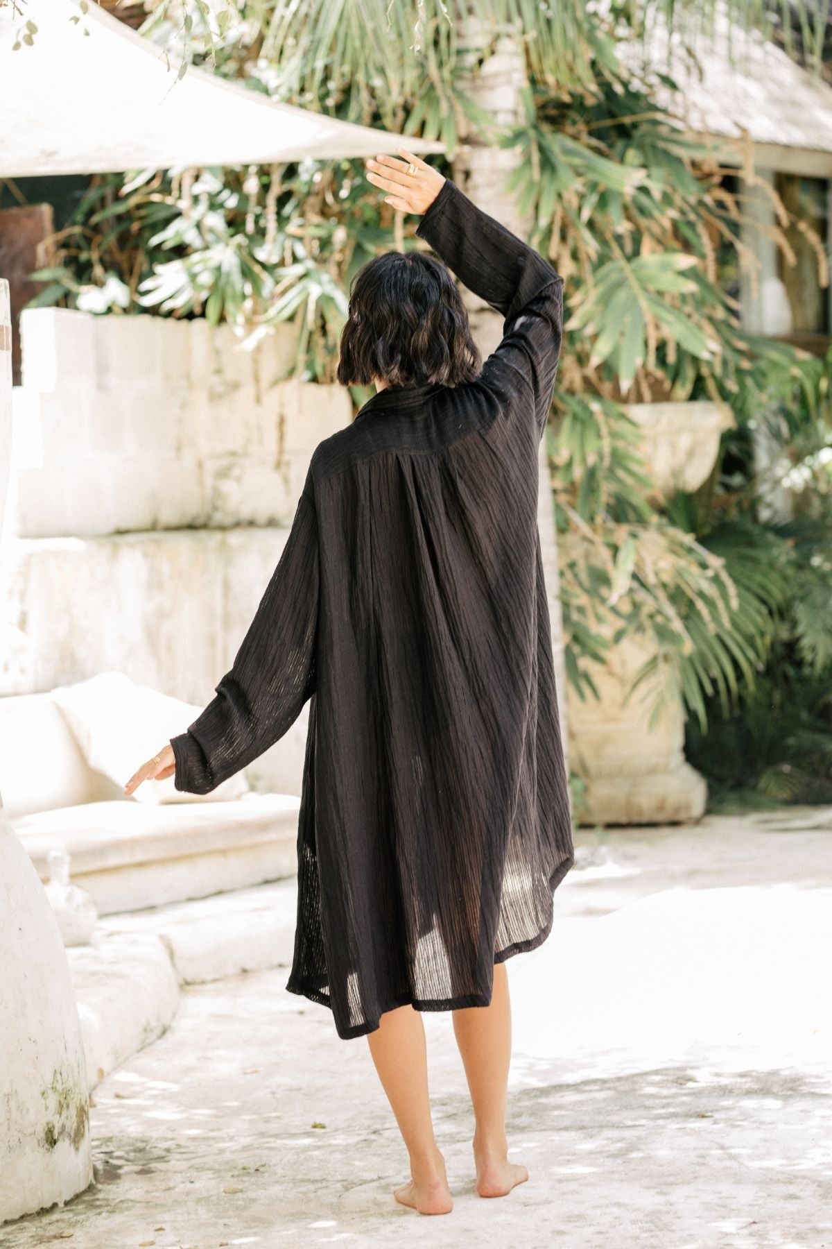 A person with short, dark hair is standing barefoot outdoors, facing away from the camera. They are wearing a loose, long-sleeved black Kundalini Gown - Short Edition (Linen/Cotton Luxe) by Myrah Penaloza. One arm is raised while the other is lowered. The background features lush greenery and stone structures.