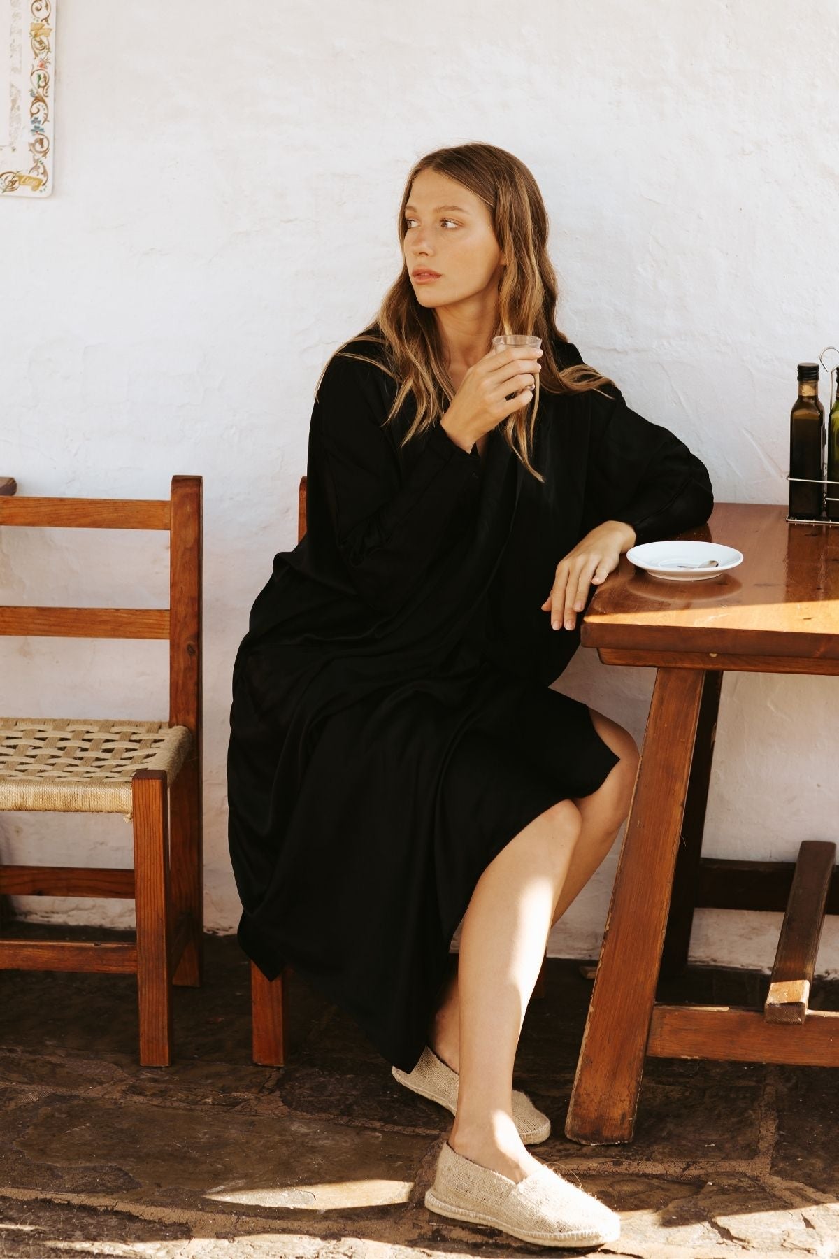 In a scene embodying intentional living, a person wearing the Myrah Penaloza Amma Gown - Dark Moon Edition sits at a wooden table against a white wall, holding a small cup. With long, wavy hair and slip-on shoes, they are surrounded by two wooden chairs, a bottle, and a small plate nearby.