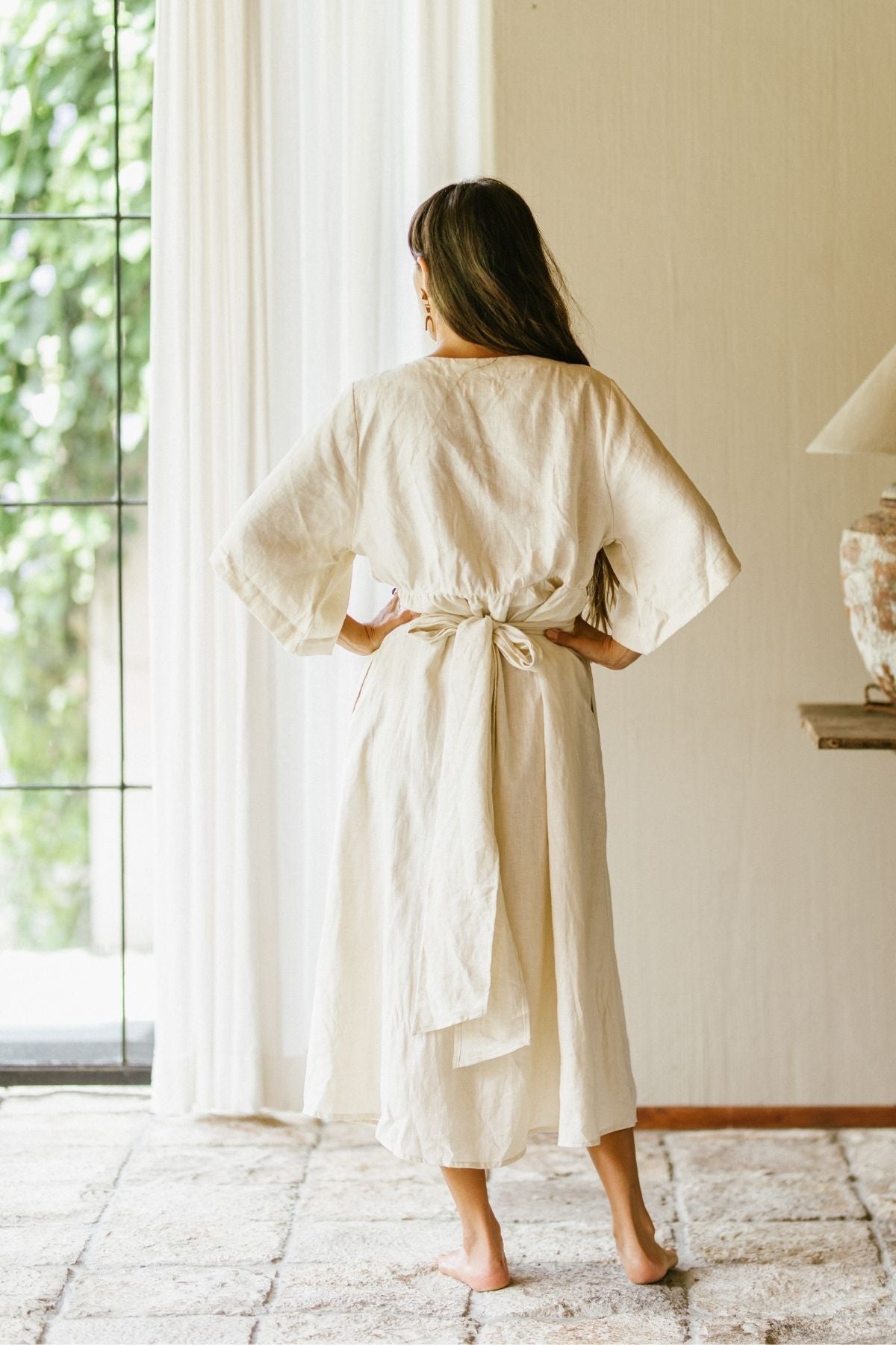 A woman stands barefoot indoors, facing away in a 100% linen Magdalena Gown, named "Moonlight," by Myrah Penaloza. The loose cream-colored robe is tied at the waist and noted for its natural durability. Soft natural light filters through a window with sheer white curtains, offering a serene view of the lush greenery outside.