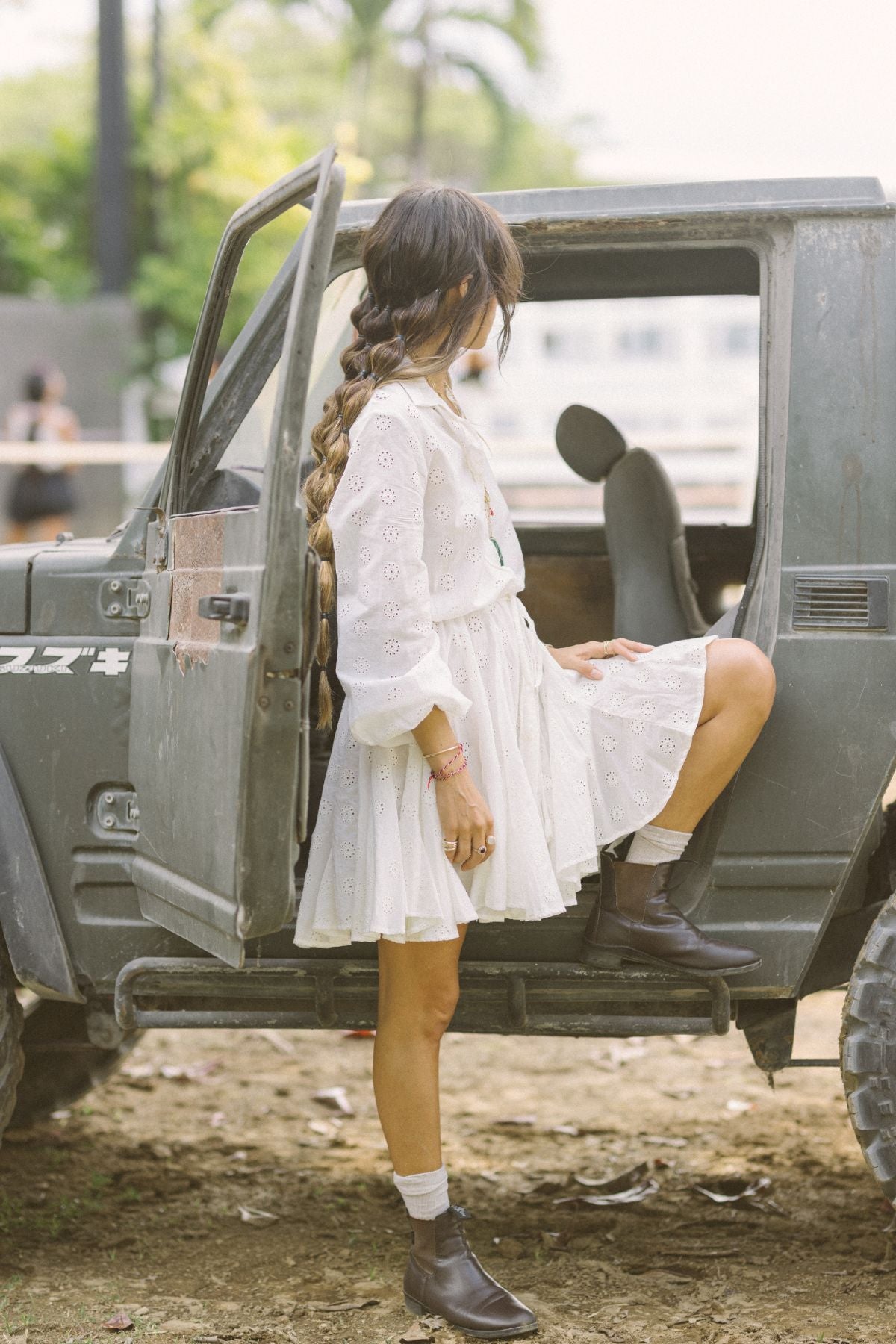 A woman with a long braid, dressed in a vintage embroidered cotton La Rosa Dress by Myrah Penaloza and brown boots, stands by an open Jeep door. She gazes away, one foot resting inside the vehicle amid the outdoor surroundings.