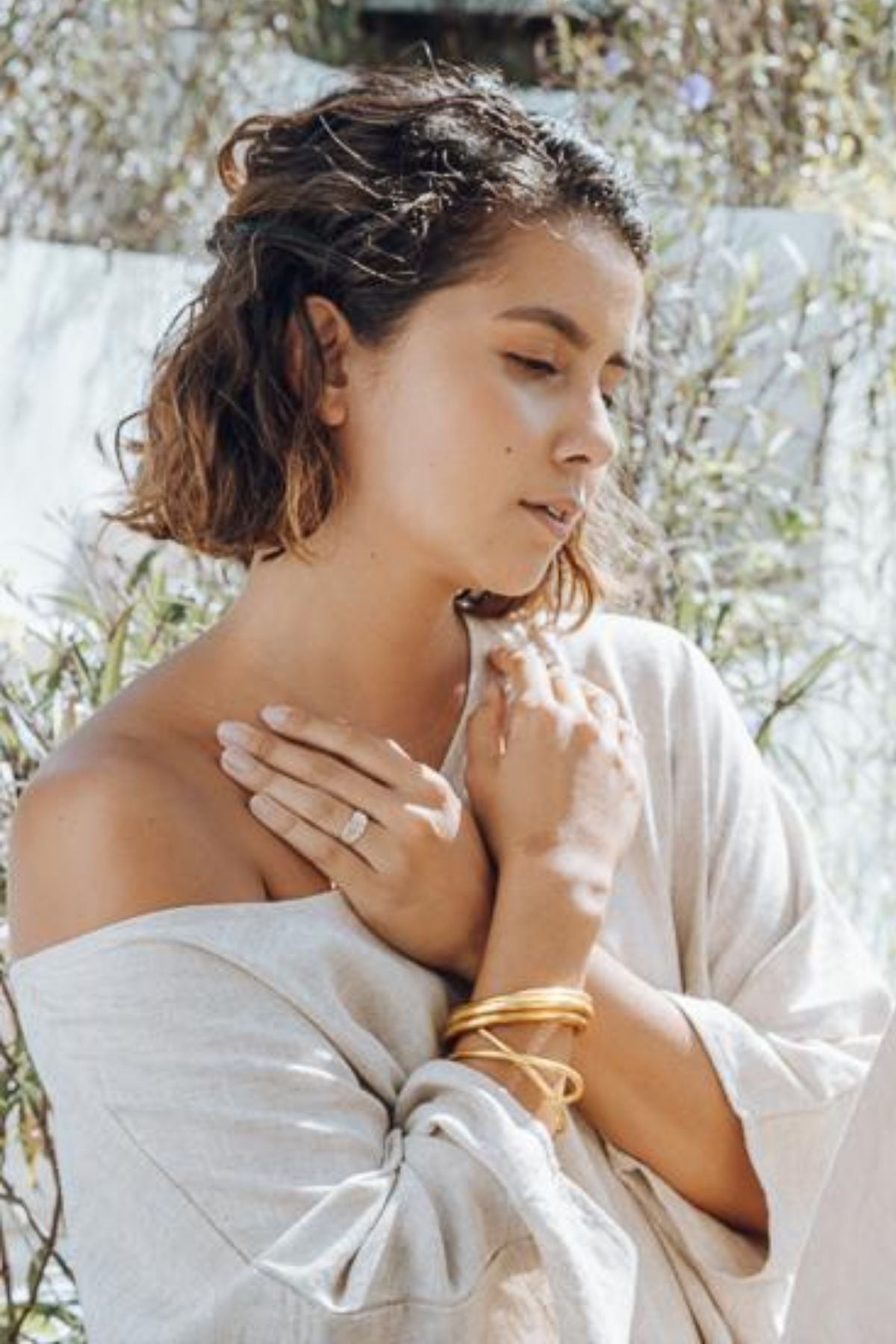 A woman with short brown hair stands outdoors with her eyes closed, wearing a light off-shoulder Butterfly Tunic by Myrah Penaloza and gold bangles. She gently places her hands near her neck, surrounded by greenery in soft sunlight.