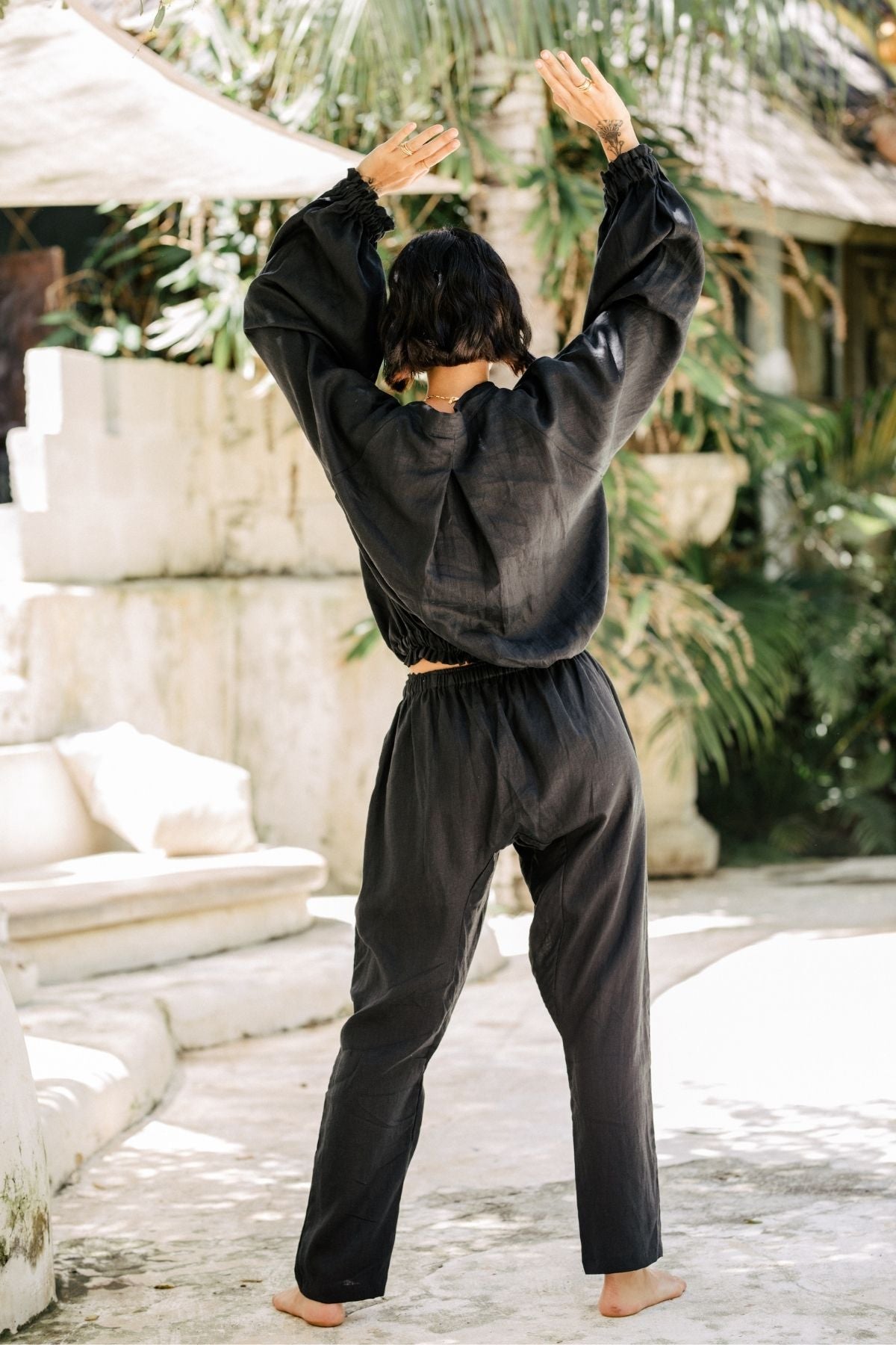 A person with short dark hair stands barefoot on a stone surface with arms raised, wearing a loose-fitting, 100% linen Frida Top in black by Myrah Penaloza. They are outdoors with greenery and a beige structure in the background, suggesting a tranquil, sustainable setting.