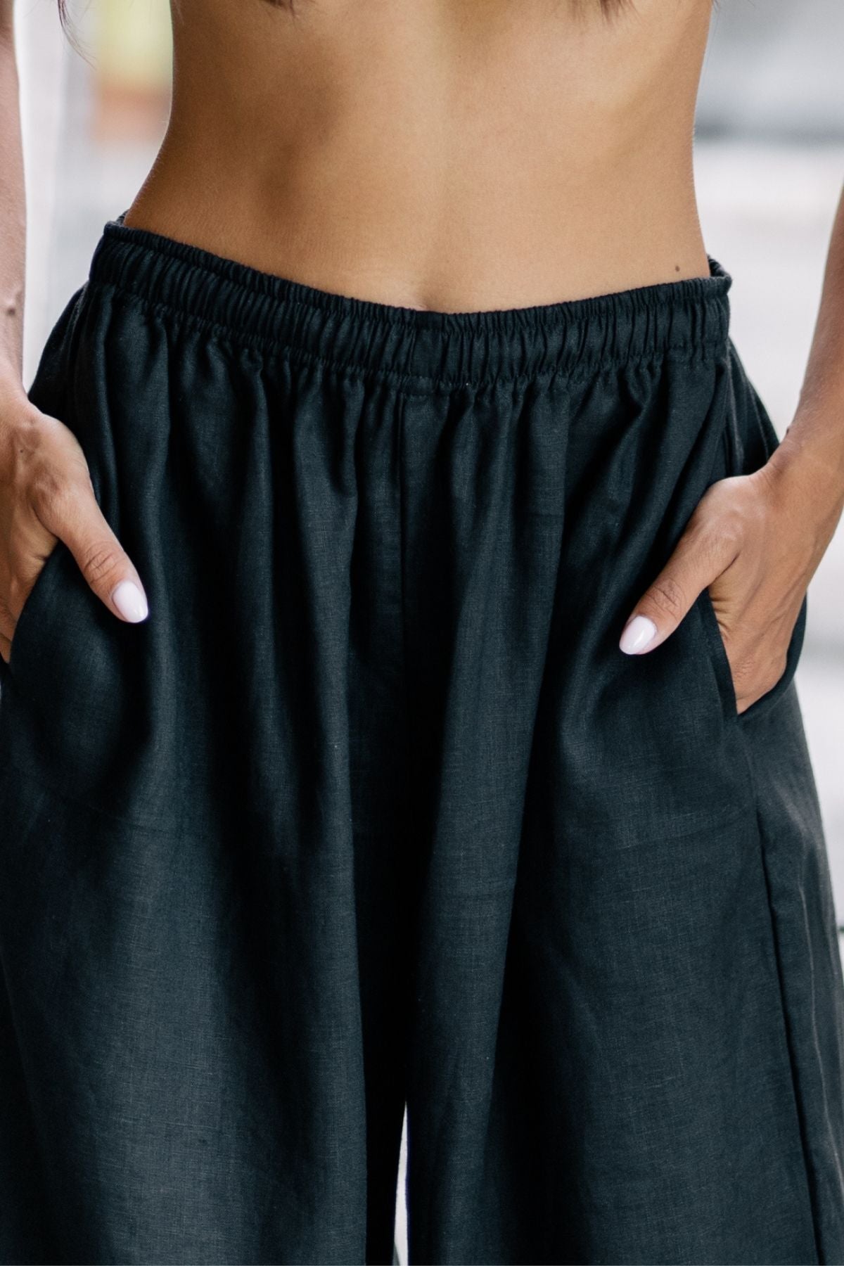 A person is shown wearing the Grace Pant by Myrah Penaloza, featuring a high-waisted design and loose-fitting wide leg in dark moon black linen with an elastic waistband. They have their hands in the pockets, showcasing a look that effortlessly blends comfort and style, making it perfect for both casual settings and office wear.