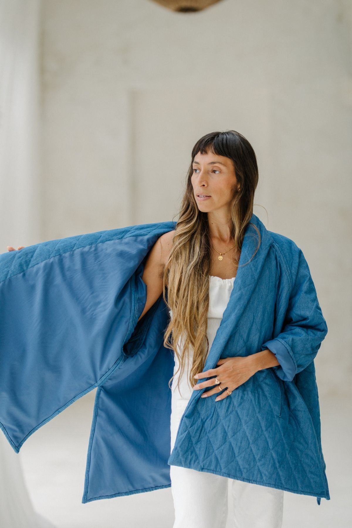 A person with long hair is wearing a Javi Quilted Jacket (Pre-Order) by Myrah Penaloza in chambray denim over a white outfit. The jacket is open, showcasing its hand-quilted inside lining. They are standing against a light, neutral background and looking off to the side.