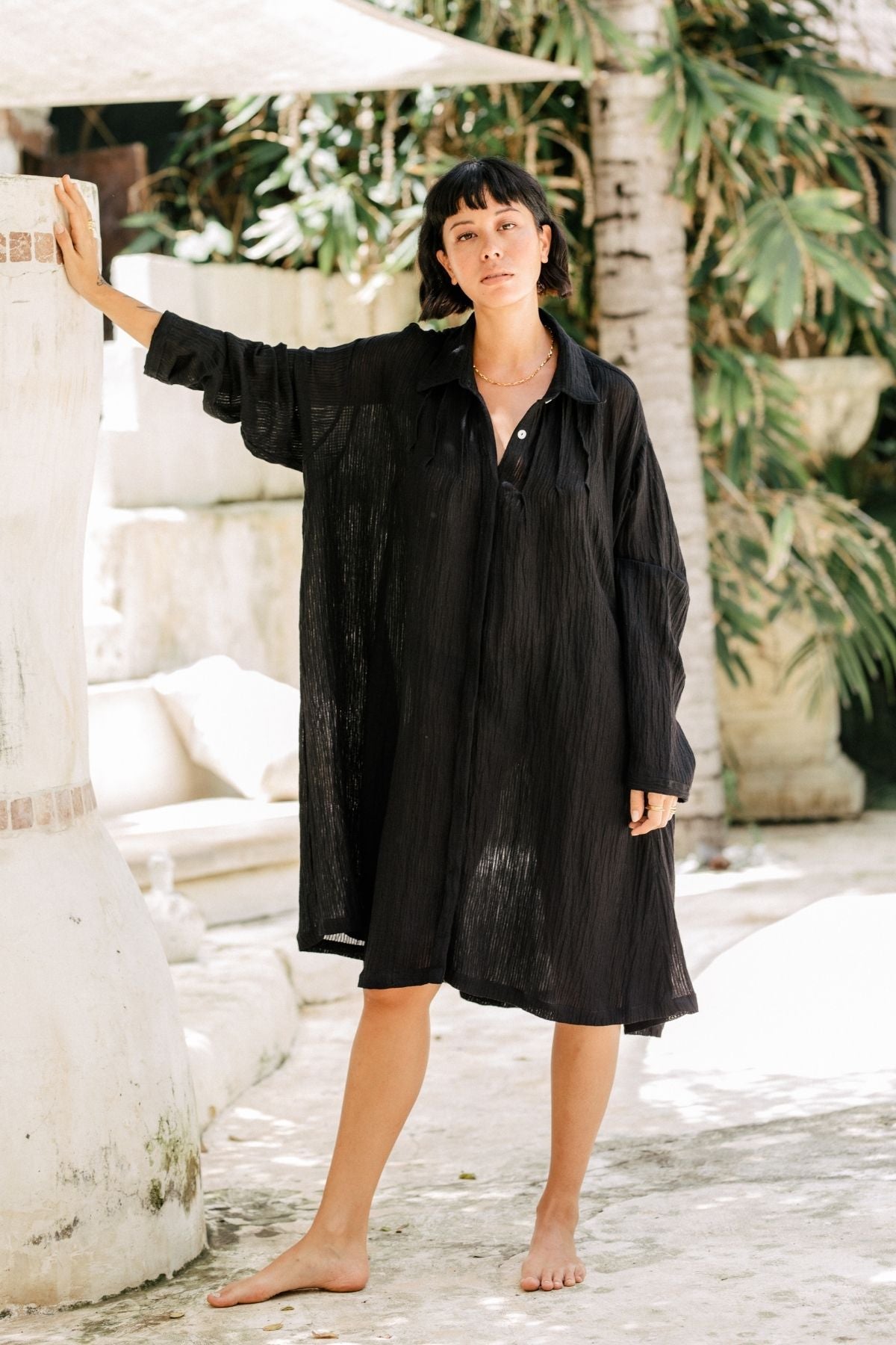 A barefoot woman stands outdoors near a white wall, wearing a Myrah Penaloza Kundalini Gown - Short Edition in black, made from Linen/Cotton Luxe fabric. She has a short, dark hairstyle and leans against the wall with her left hand. Lush green plants and sunlight creating shadows in the background complete this chic look.