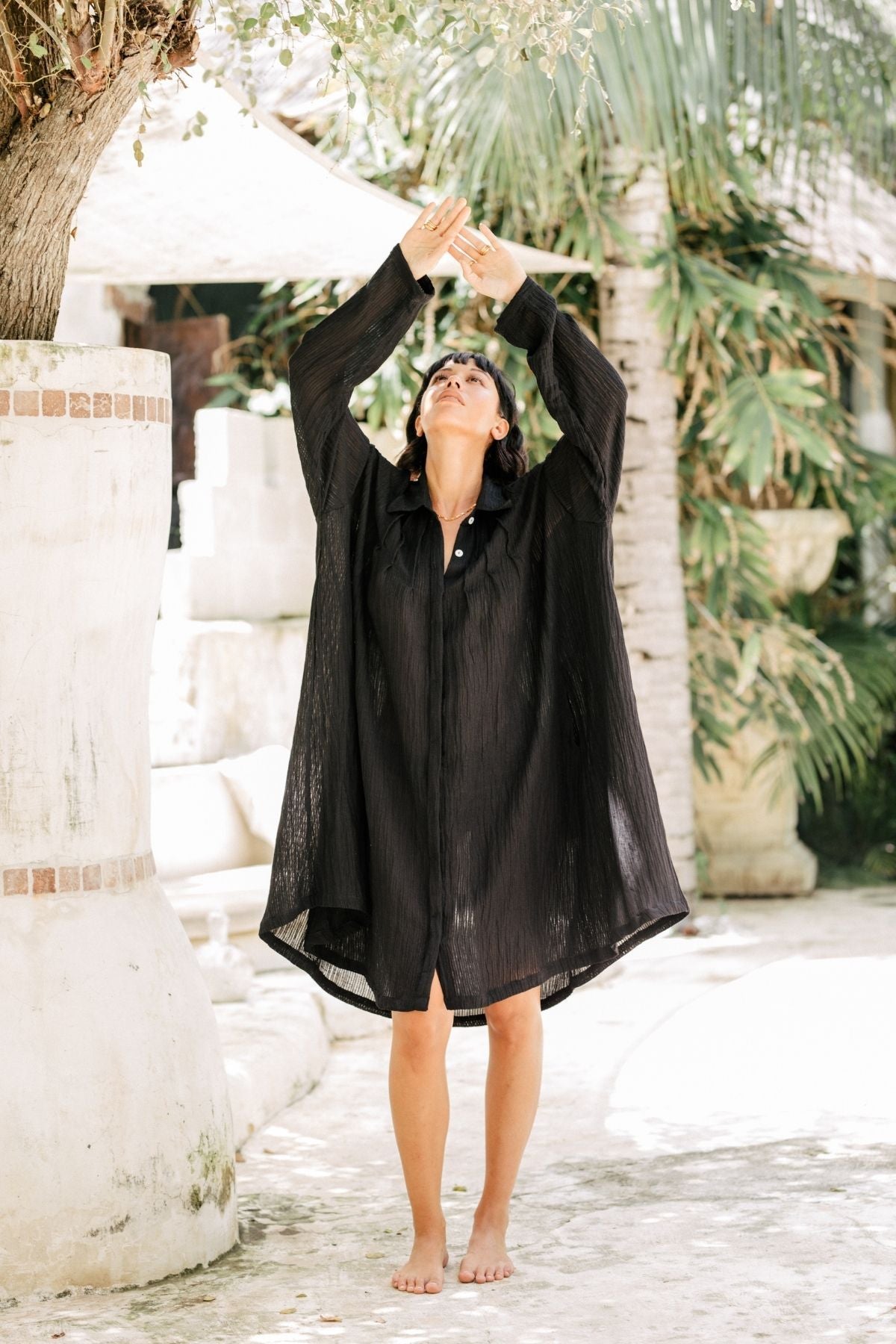 A person stands barefoot on a pathway, stretching their arms up towards a tree. They are wearing a loose-fitting black Kundalini Gown - Short Edition (Linen/Cotton Luxe) from Myrah Penaloza and have their eyes closed. Tropical plants and stone structures surround the area, creating a serene outdoor environment.