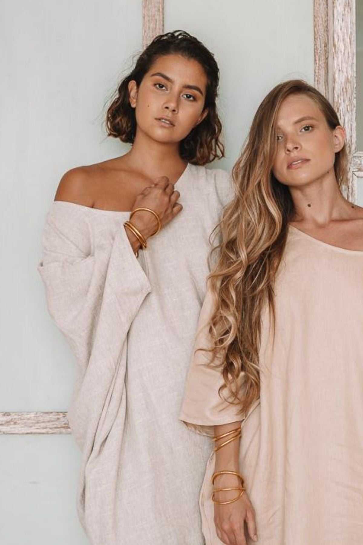 Two women stand side by side against a light-colored wall, both dressed in elegant Butterfly Tunics from Myrah Penaloza in natural tones and accessorized with gold bangles. The woman on the left sports short, wavy hair, while the one on the right showcases long waves. Both display neutral expressions.