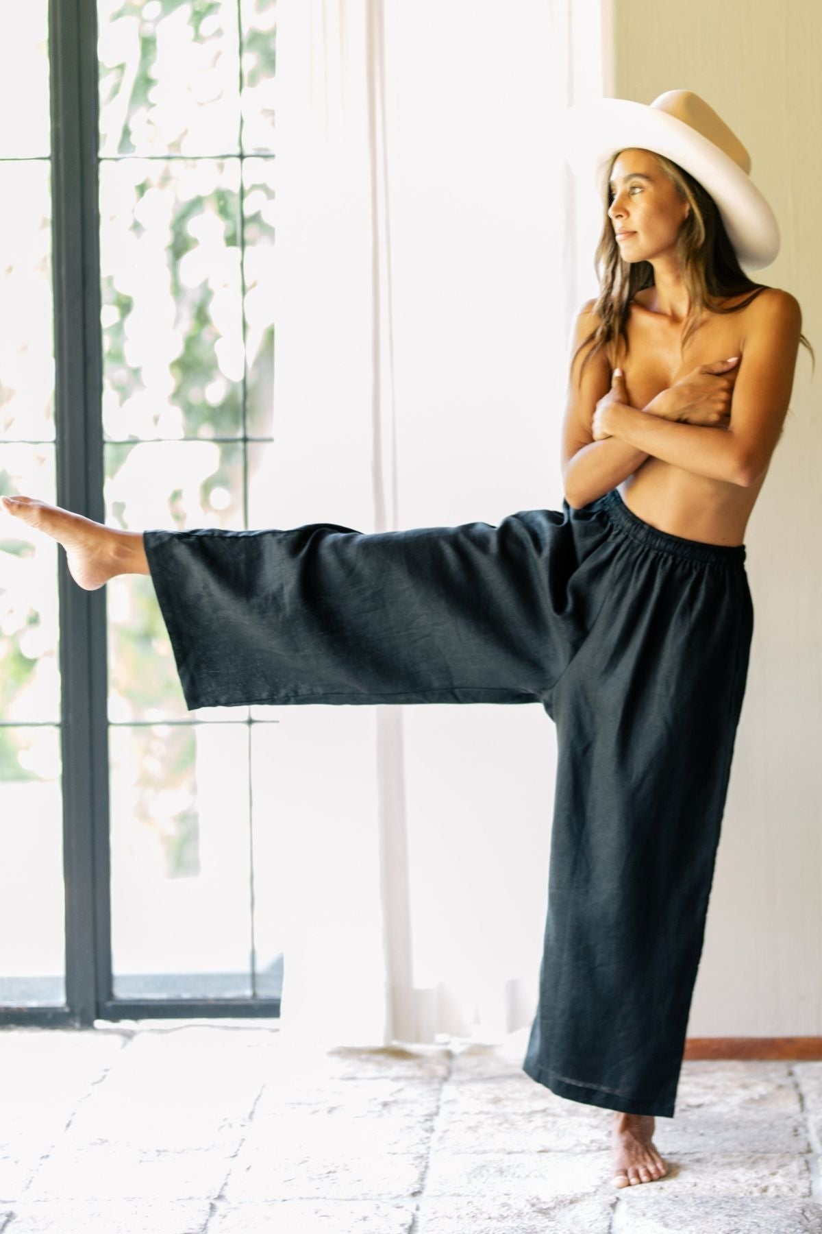 A woman stands barefoot indoors, wearing a wide-brimmed hat and the Grace Pant in dark moon black, crafted from 100% linen by Myrah Penaloza. She crosses her arms and extends one leg sideways to maintain her balance as sunlight filters through a large window, casting soft light on her and the room.