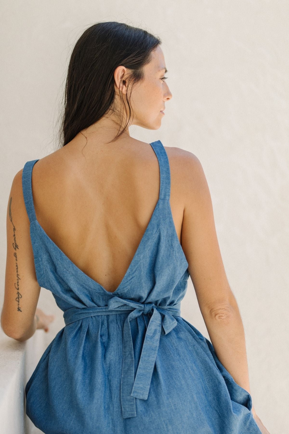 A woman with long dark hair is wearing the Denim Stonewashed Kundalini Playsuit by Myrah Penaloza, featuring a sleeveless design and a low back with a tied belt. She stands sideways against a light-colored wall, looking forward, while a tattoo enhances her right arm, adding an intriguing element to her relaxed yet stylish ensemble.