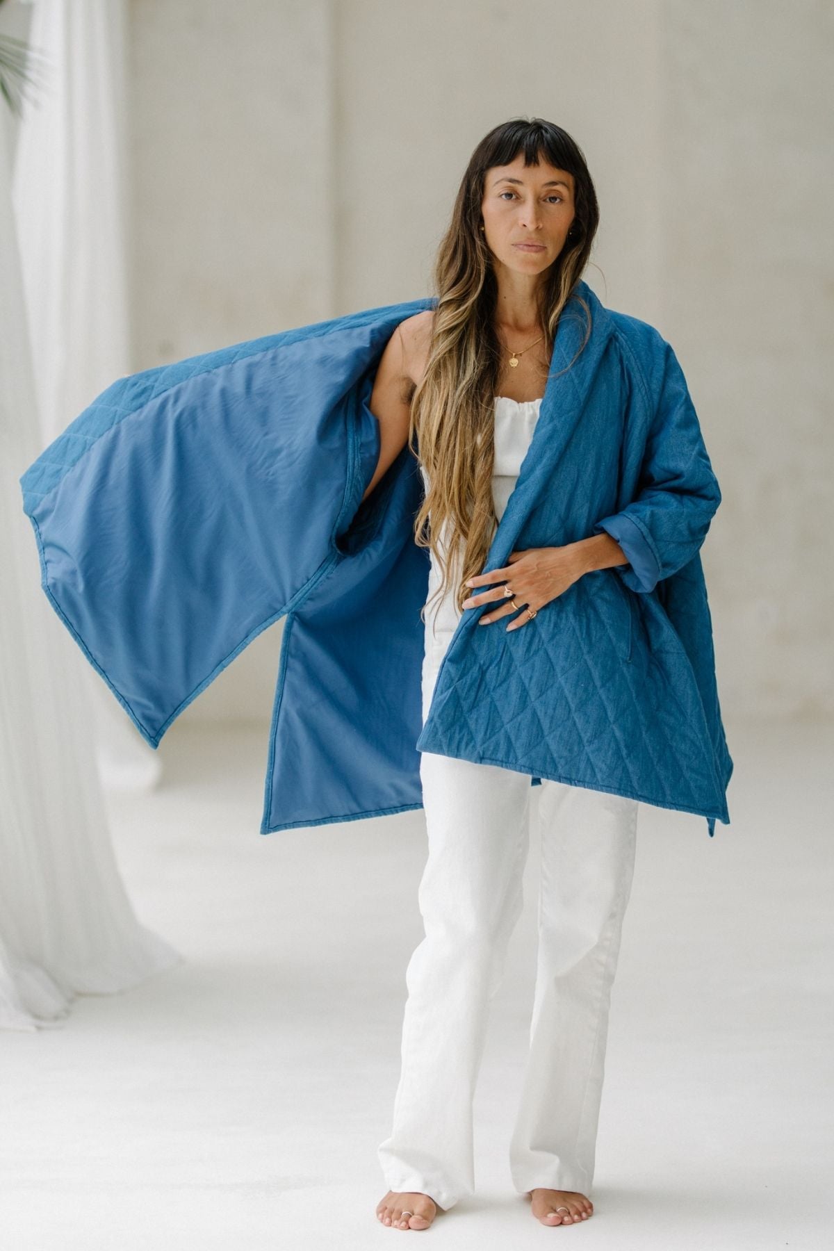 A person with long hair stands barefoot on a white floor, dressed in white top and pants. They hold open the Myrah Penaloza Javi Quilted Jacket (Pre-Order), showcasing its hand-quilted inner lining. The background is light-colored, adorned with draped white fabric, creating a minimalist aesthetic.